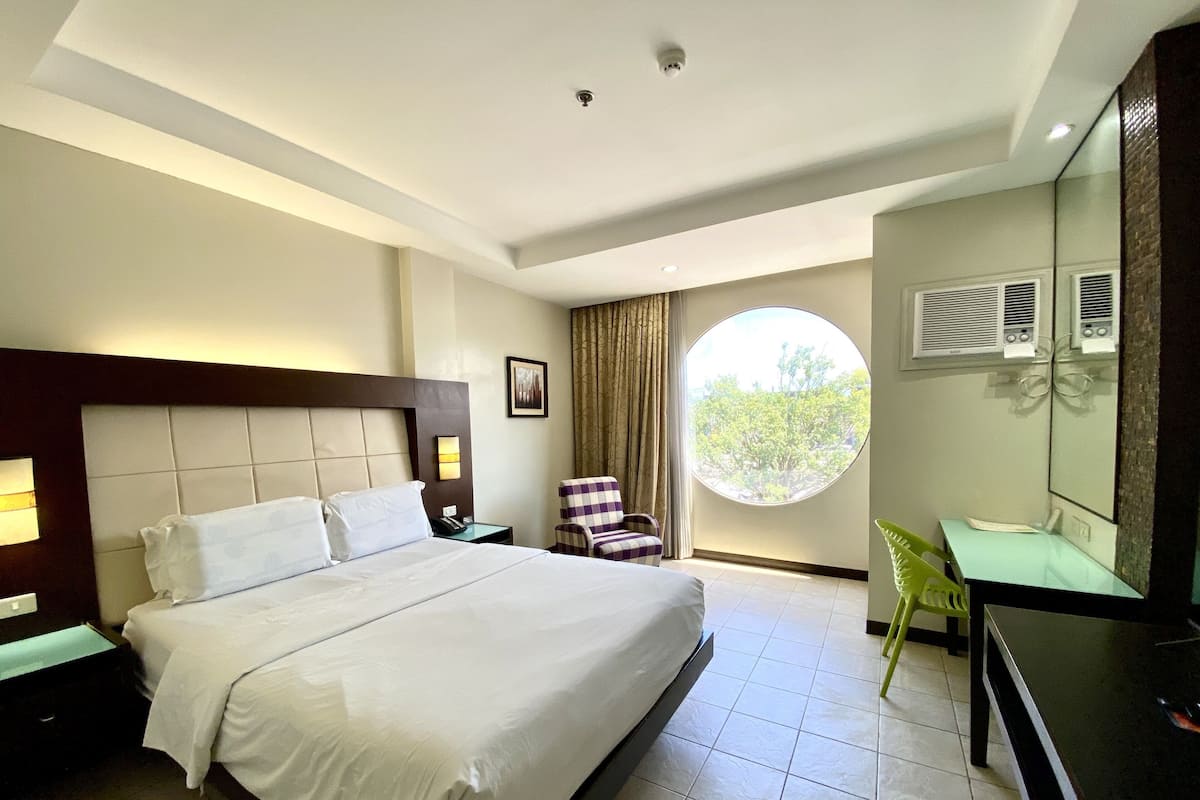 Quarto de hotel com uma cama grande, uma escrivaninha com cadeira, um janelão com vista e um espelho na parede.