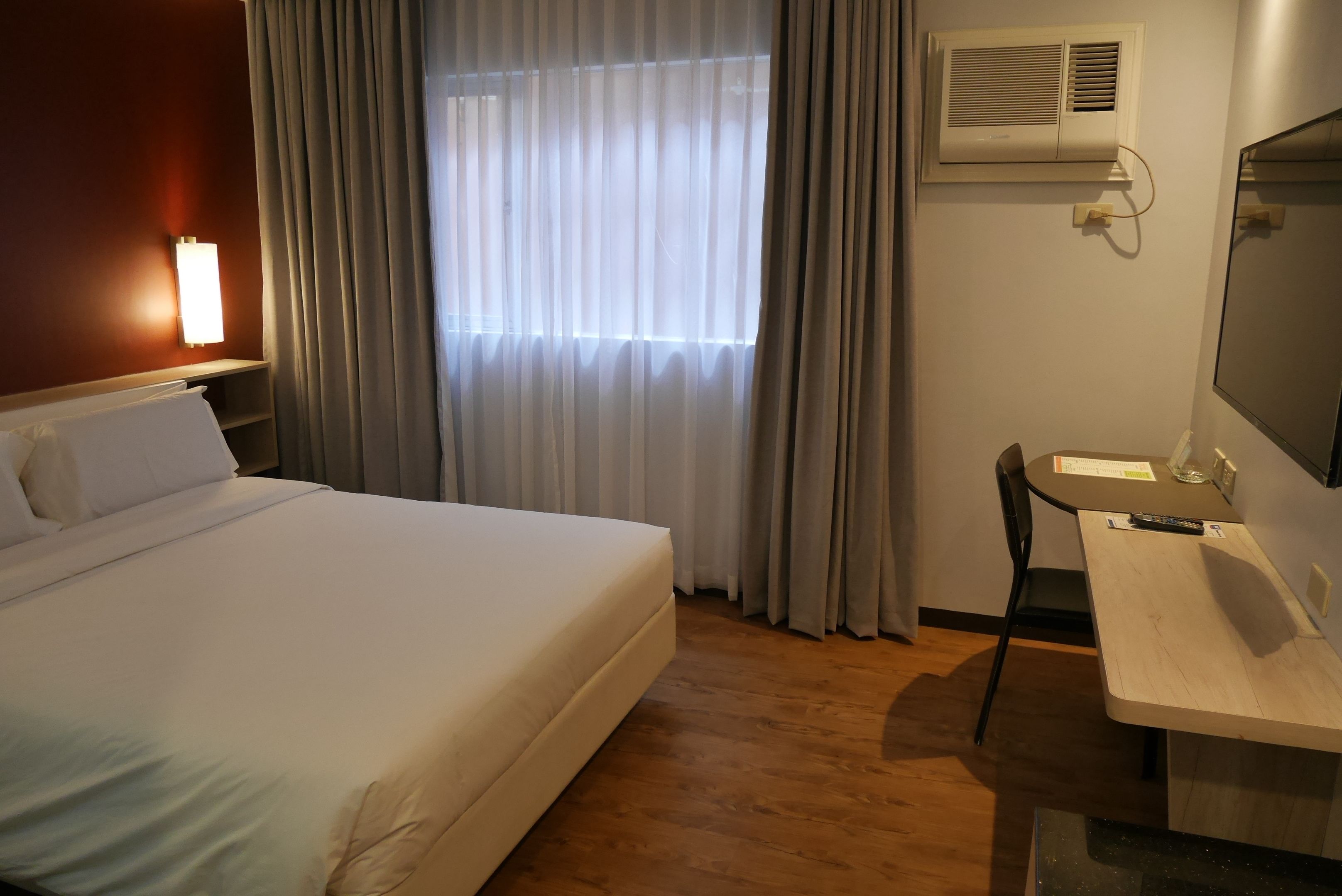 Hotelkamer met een groot bed, een bureau, een stoel, een raam met gordijnen, een airconditioner en een wandlamp.