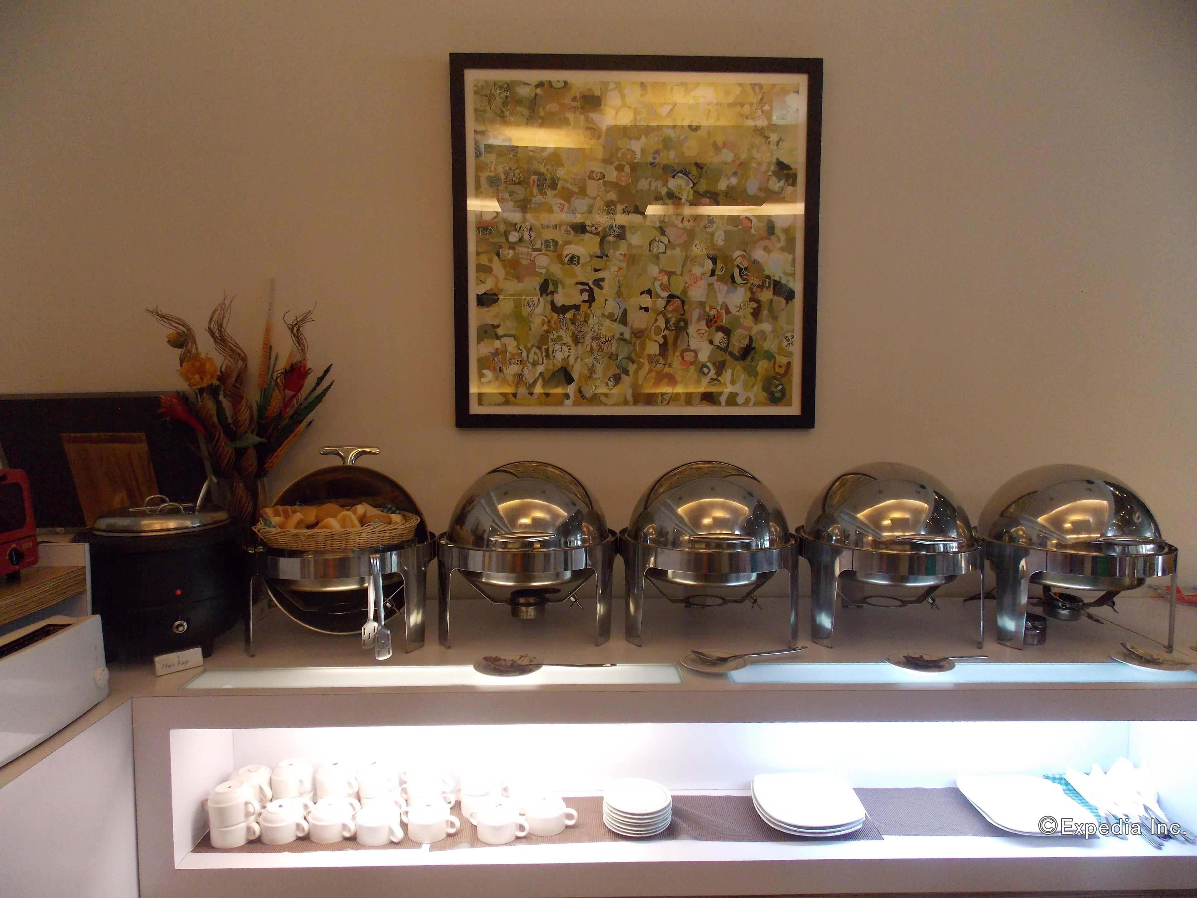 A buffet setup with covered food containers, a framed artwork above, and a display of white cups and plates below.
