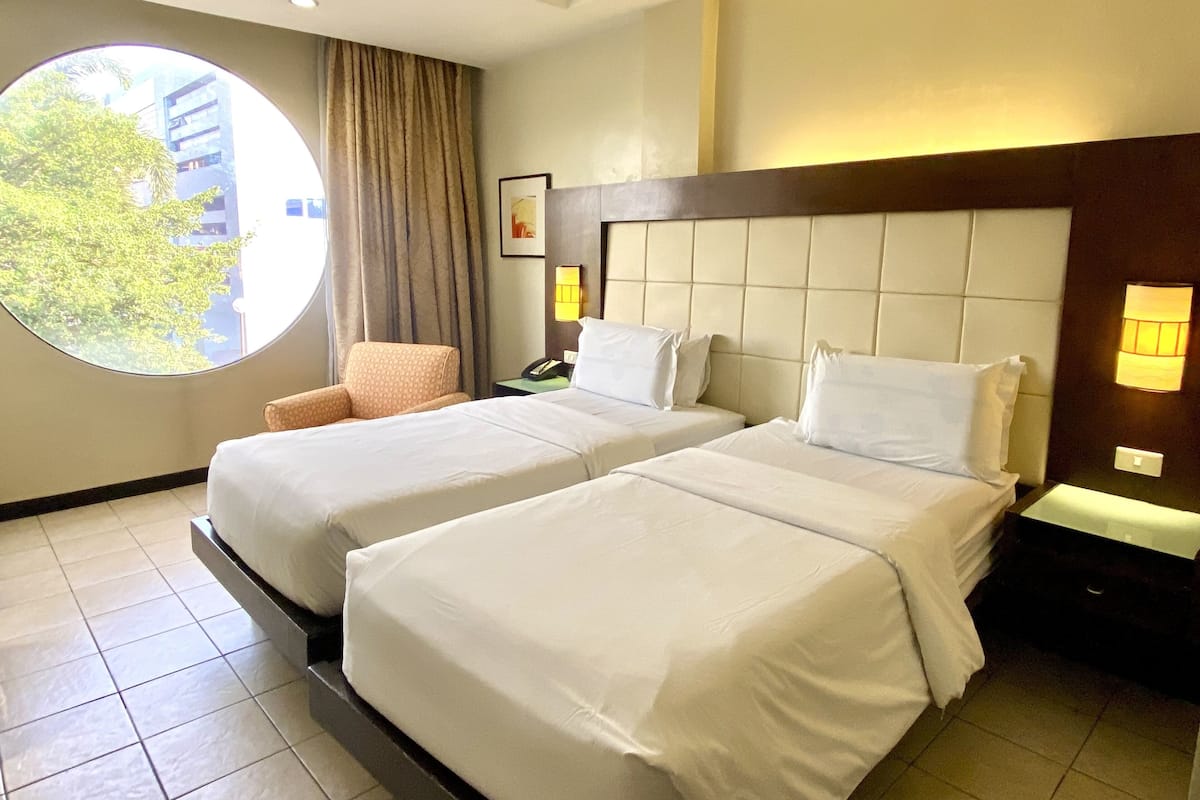 Quarto de hotel com duas camas, uma janela redonda, uma cadeira e uma mesa de cabeceira com telefone.