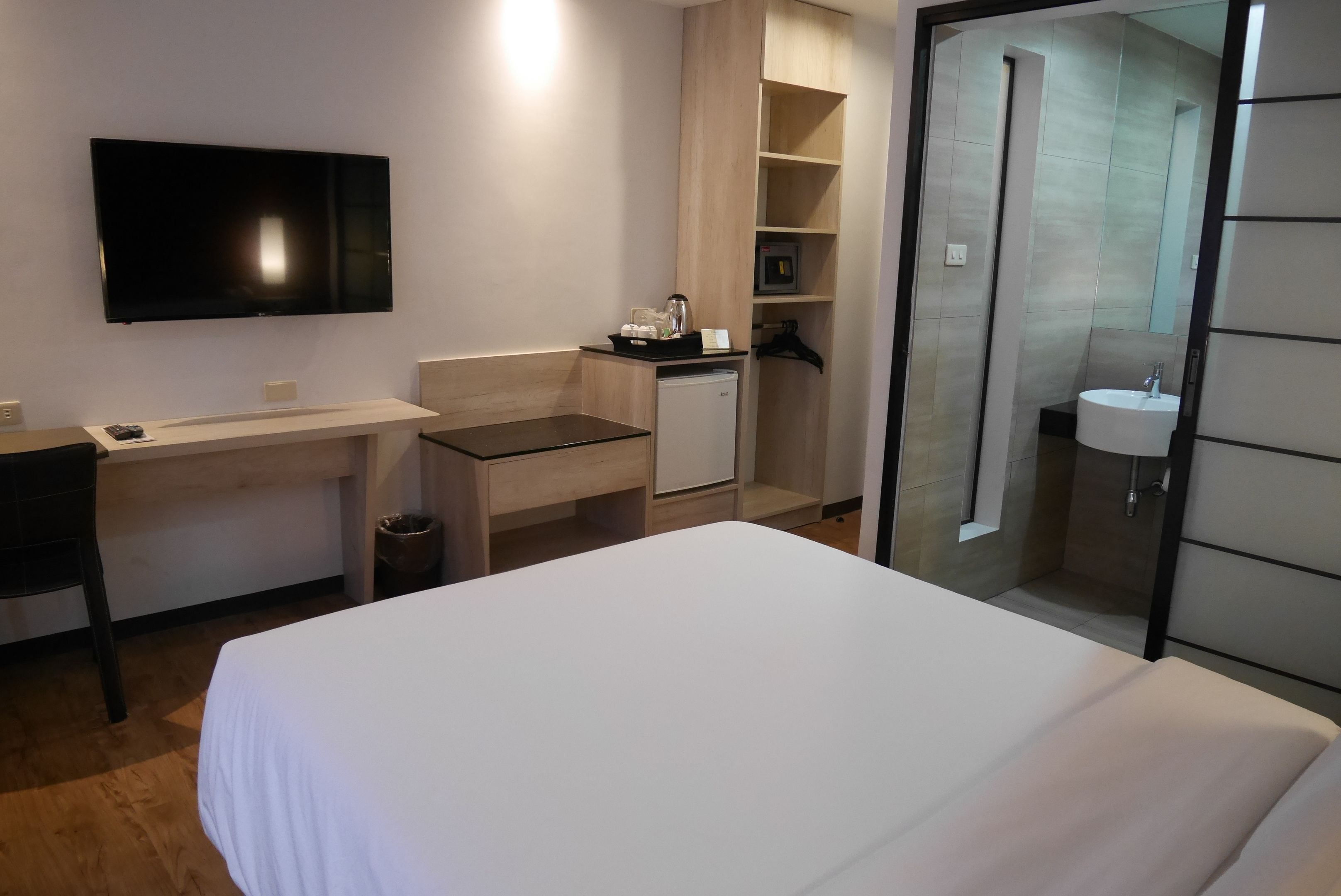 Habitación de hotel con cama, escritorio, televisor, refrigerador pequeño y zona de cocina, además de baño con lavamanos y espejo.