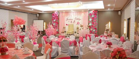 A banquet hall with round tables covered in white tablecloths and pink sashes, a stage with floral decorations, and a white screen in the background.