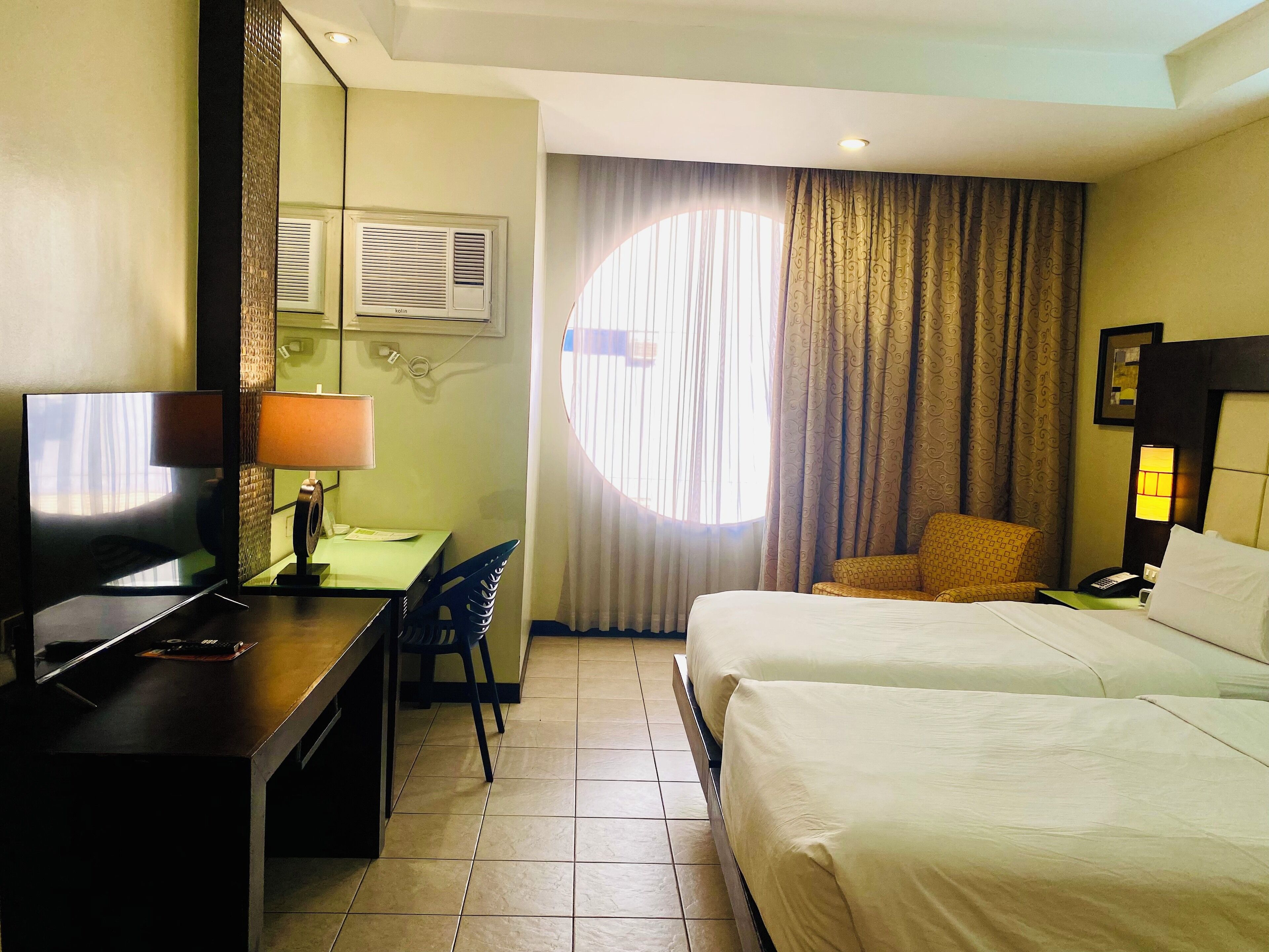 Una habitación de hotel con una cama, un escritorio con lámpara, una silla, un televisor y una ventana con cortinas.