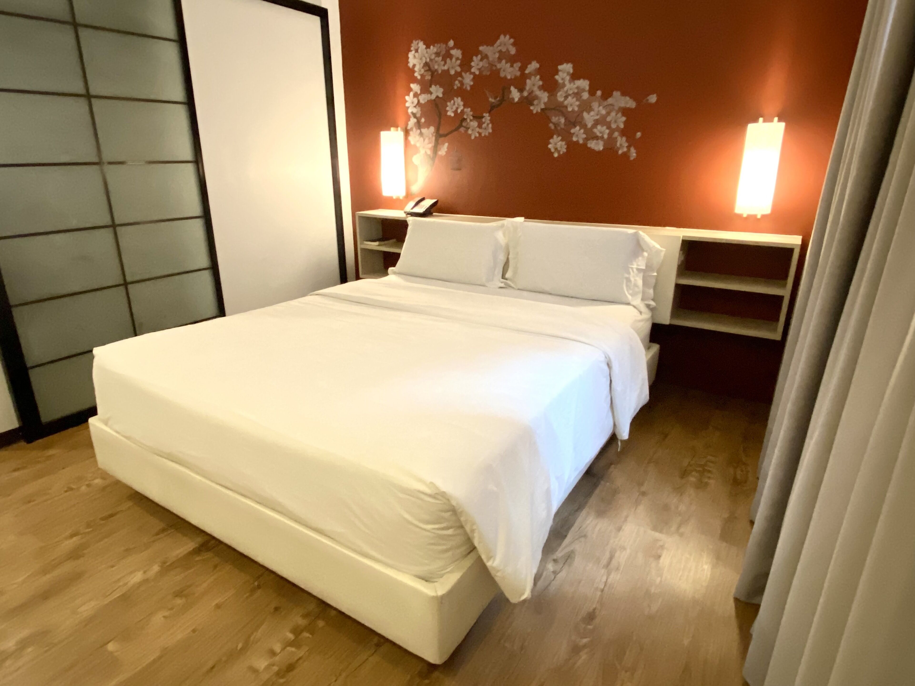 A neatly made bed in a room with a wooden floor, a wall-mounted shelf with a telephone, and a decorative wall piece.