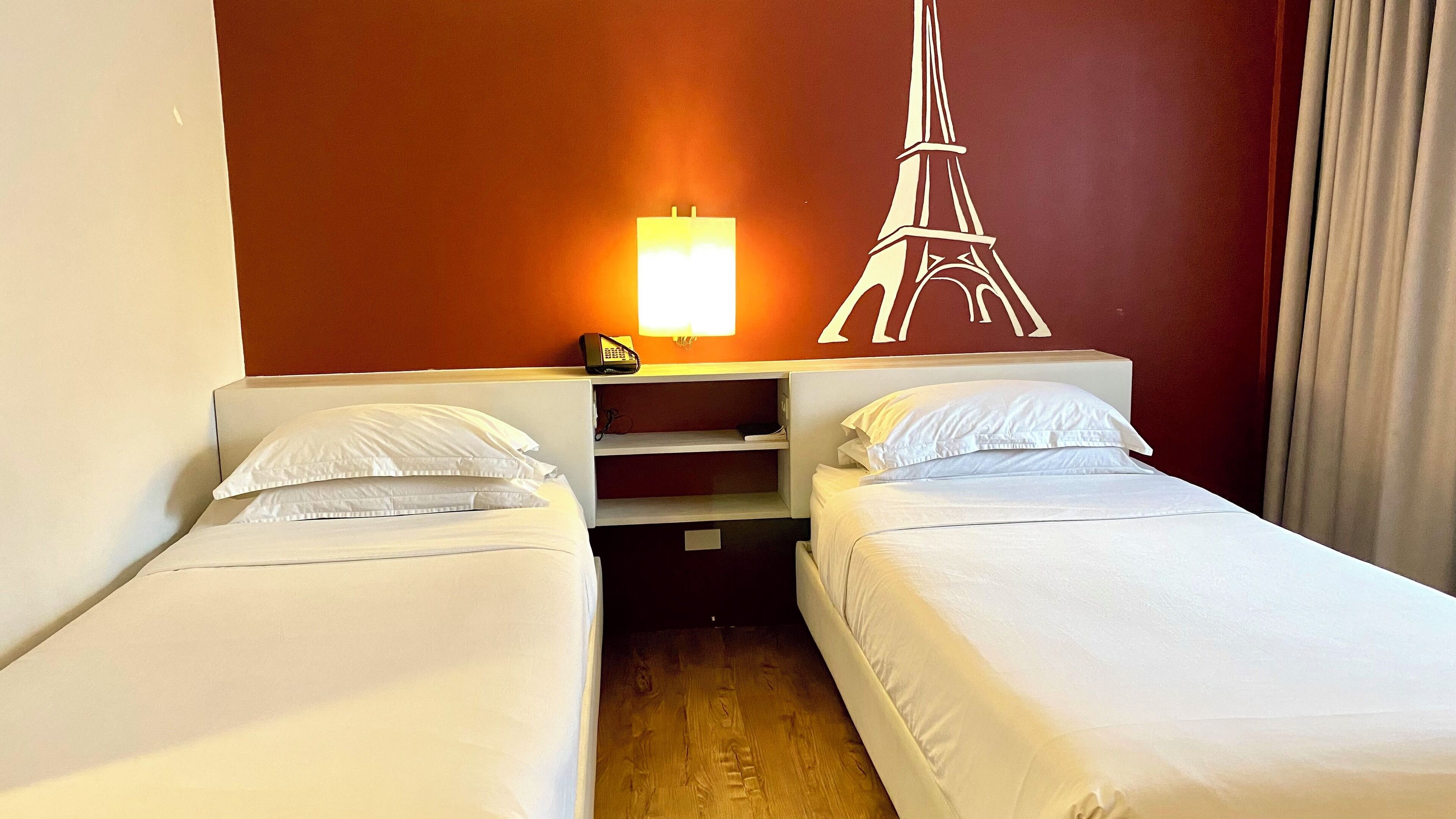 Ein Hotelzimmer mit zwei Einzelbetten, einem Nachttisch mit Lampe und einer Wanddekoration mit dem Eiffelturm.