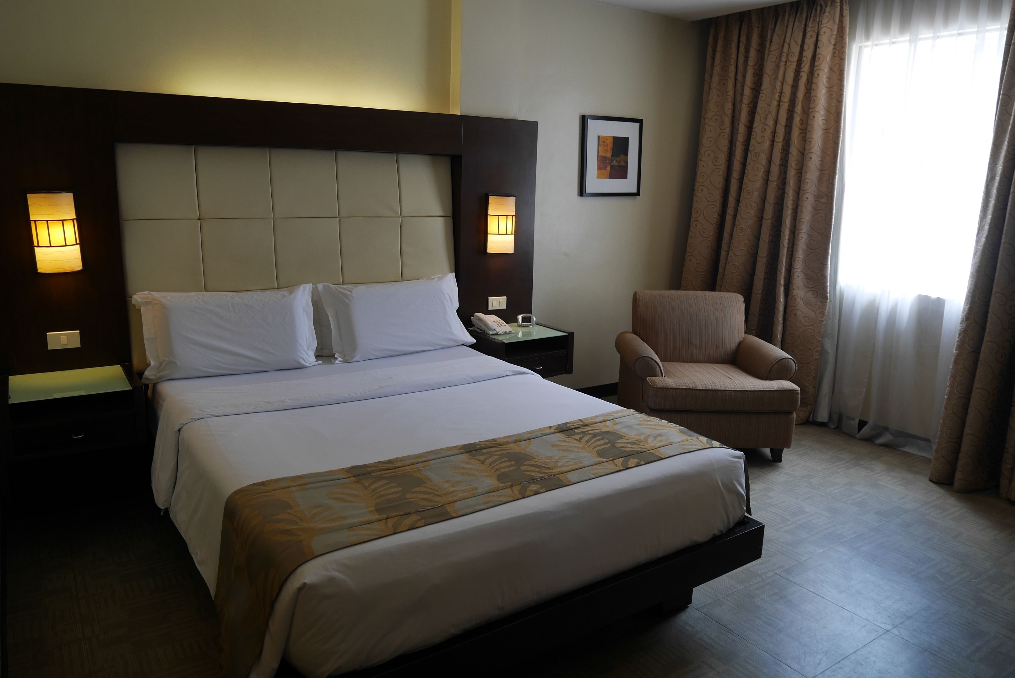 A hotel room with a large bed, a chair, a nightstand, a lamp, and a window with curtains.
