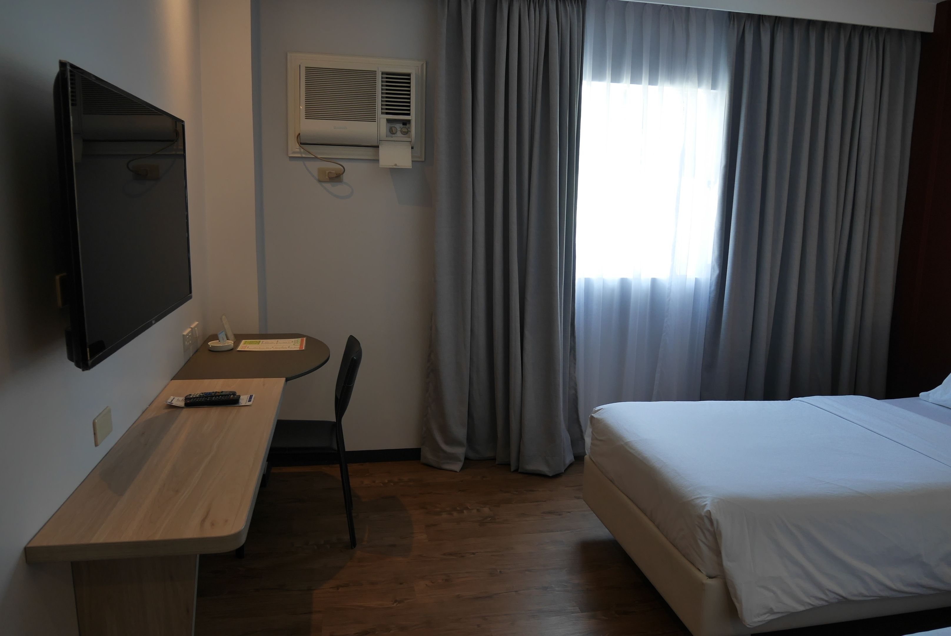 A hotel room with a bed, a desk with a chair, a TV, and an air conditioning unit.