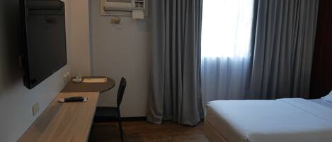 A hotel room with a bed, a desk with a chair, a TV, and an air conditioning unit.