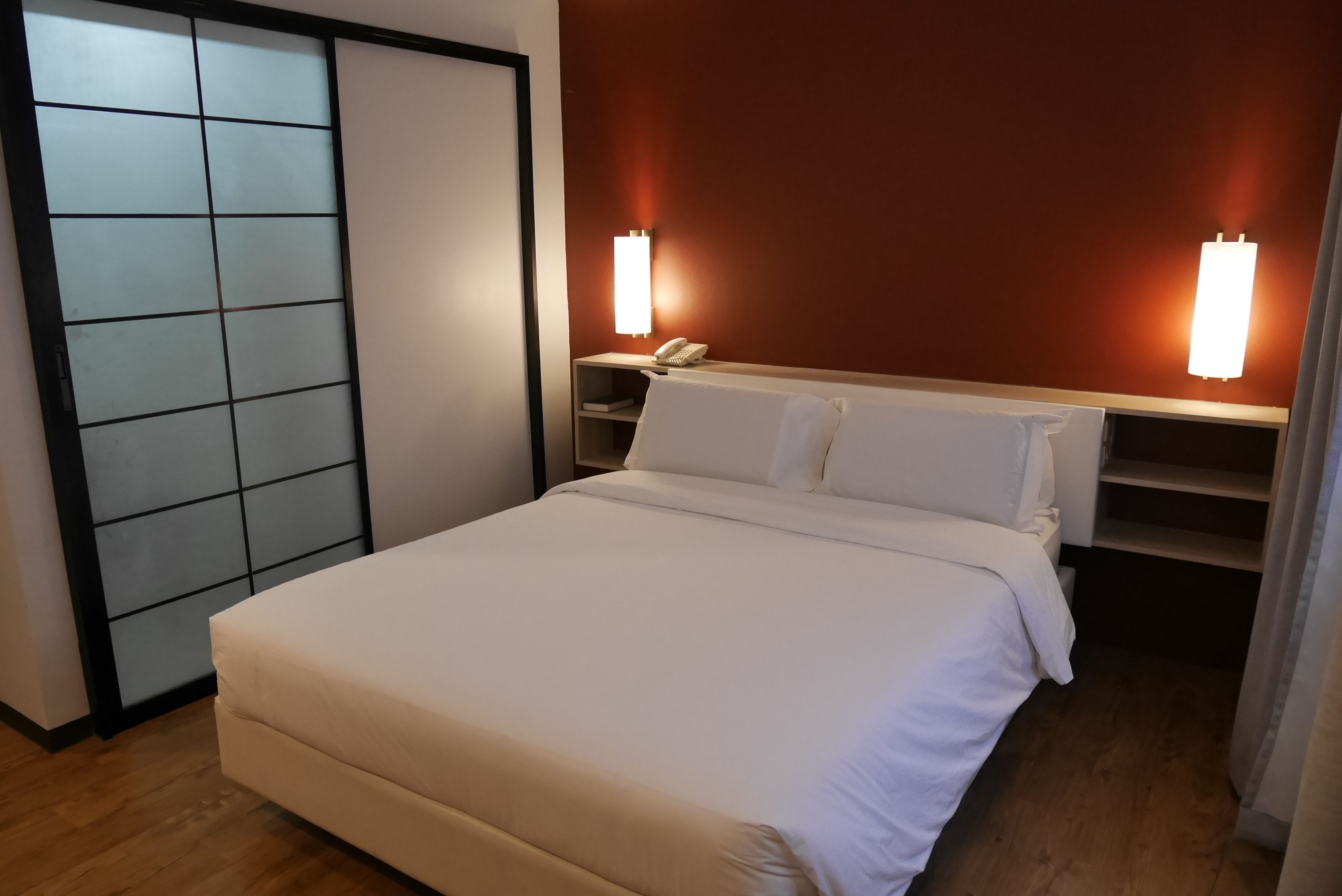 A neatly made bed in a hotel room with a wooden floor, a sliding door, and two bedside lamps.