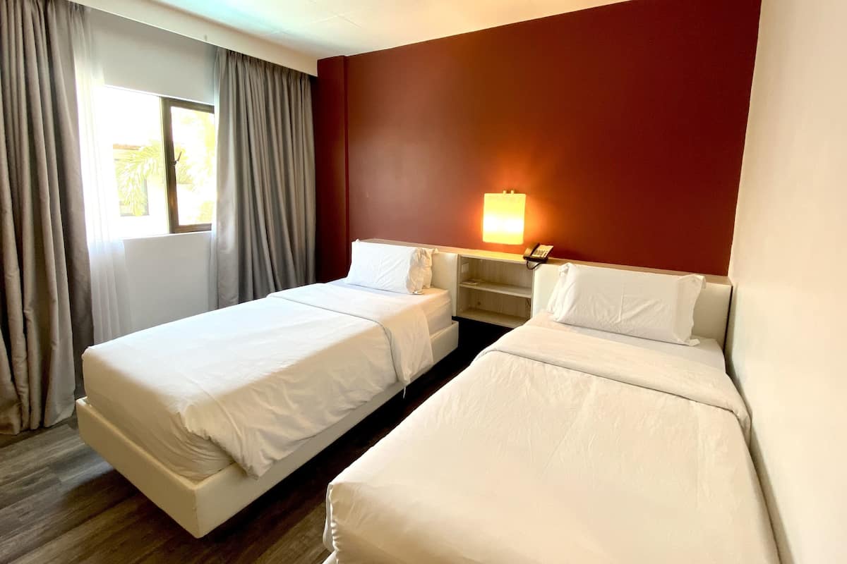 Quarto de hotel com duas camas de solteiro, mesa de cabeceira com abajur, janela com cortinas e uma parede com detalhes em vermelho.