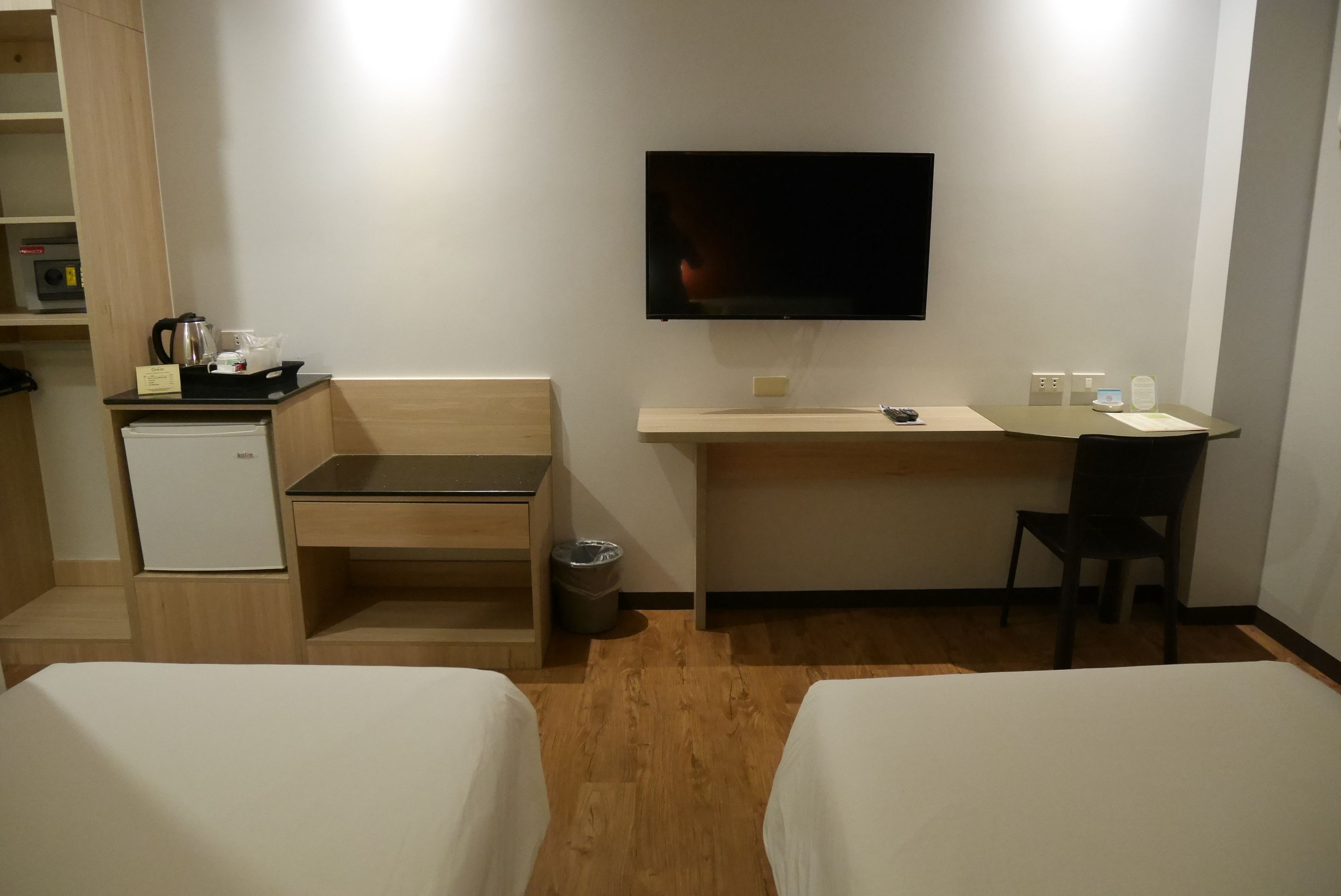 A hotel room with two beds, a desk, a television, a small refrigerator, and a trash bin.