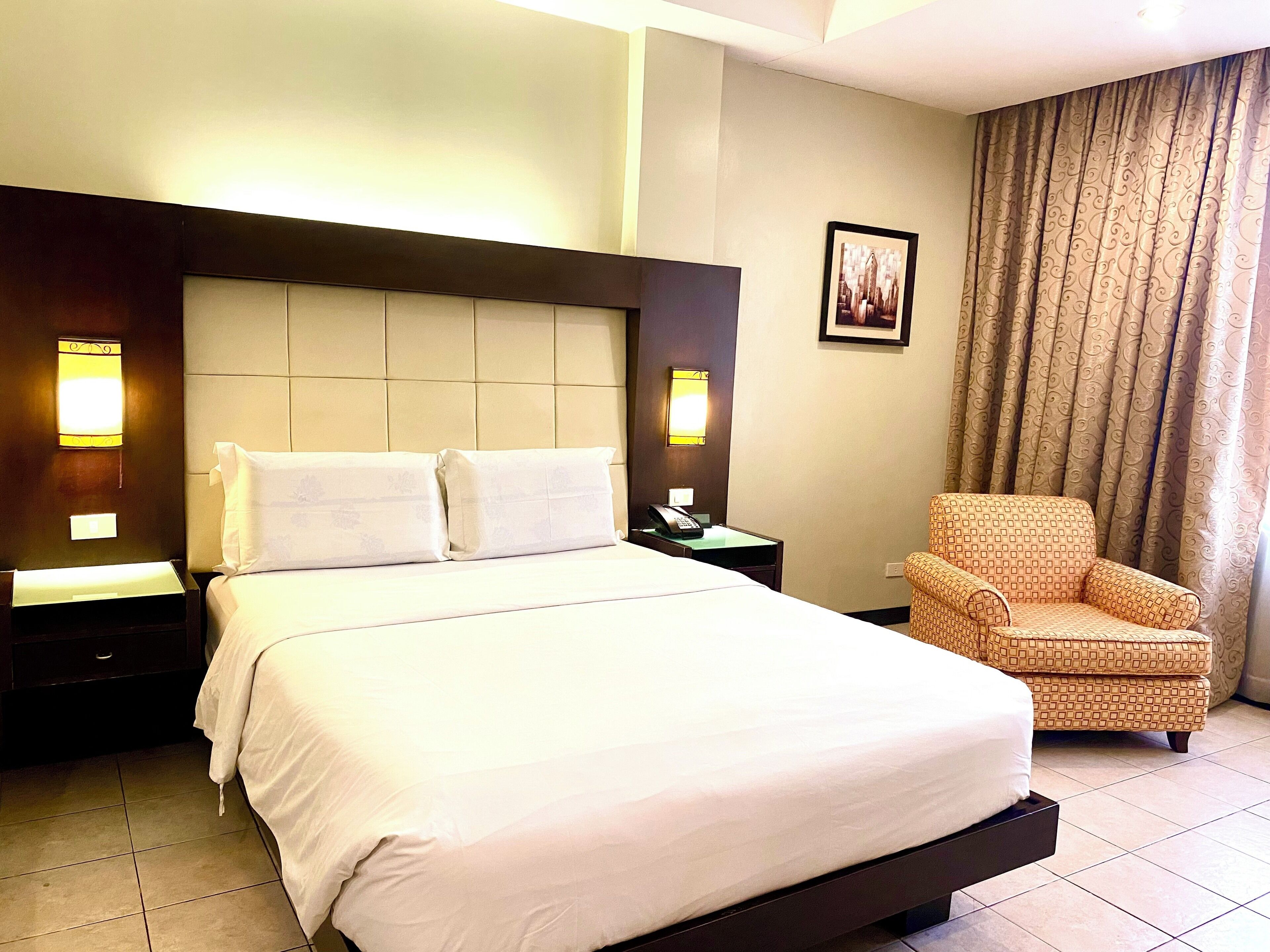 A hotel room with a large bed, a nightstand with a telephone, a chair, and a picture on the wall.