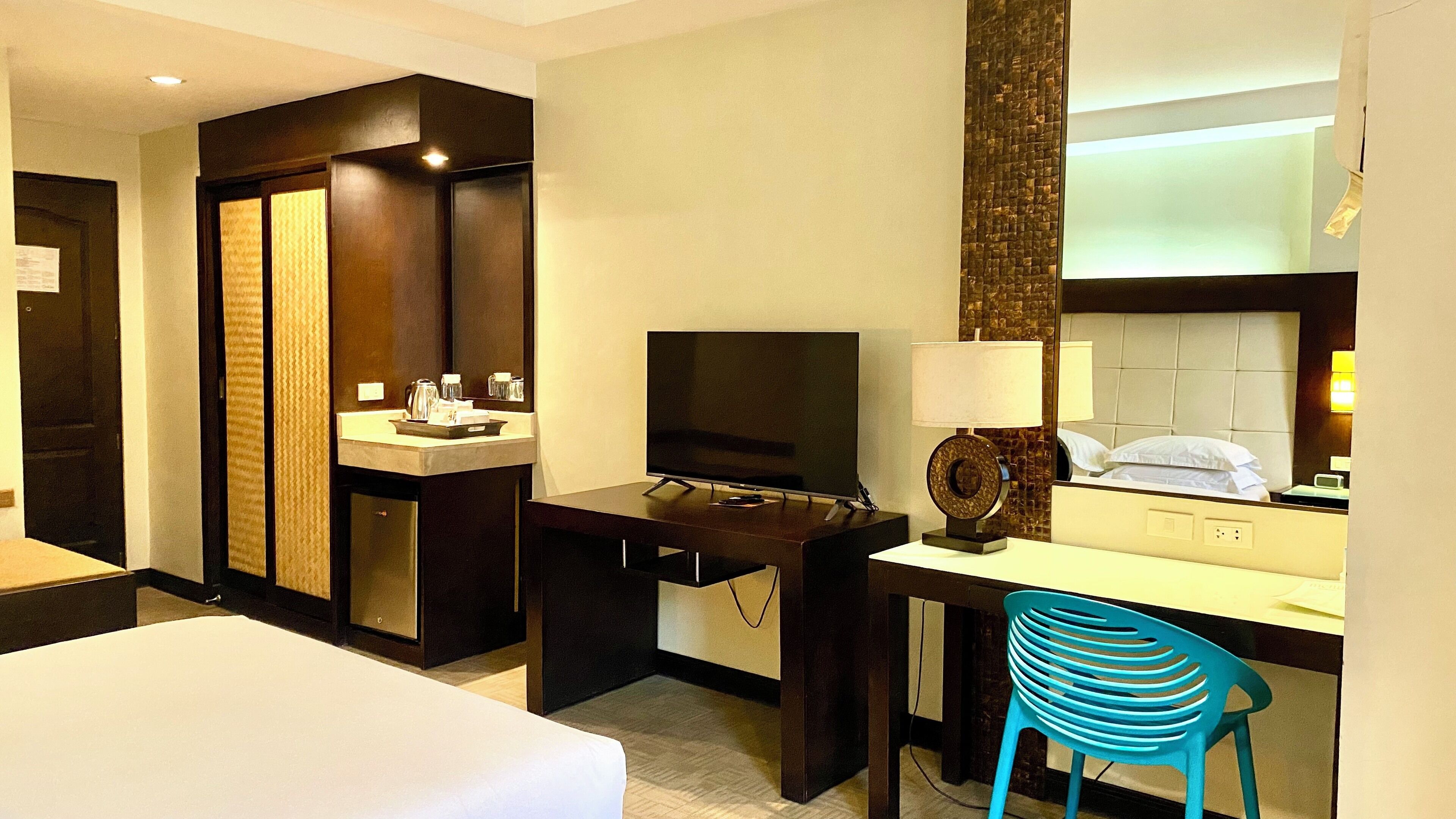 A hotel room with a bed, a desk with a television, a mirror, a bathroom with a sink and a toilet, and a blue chair.