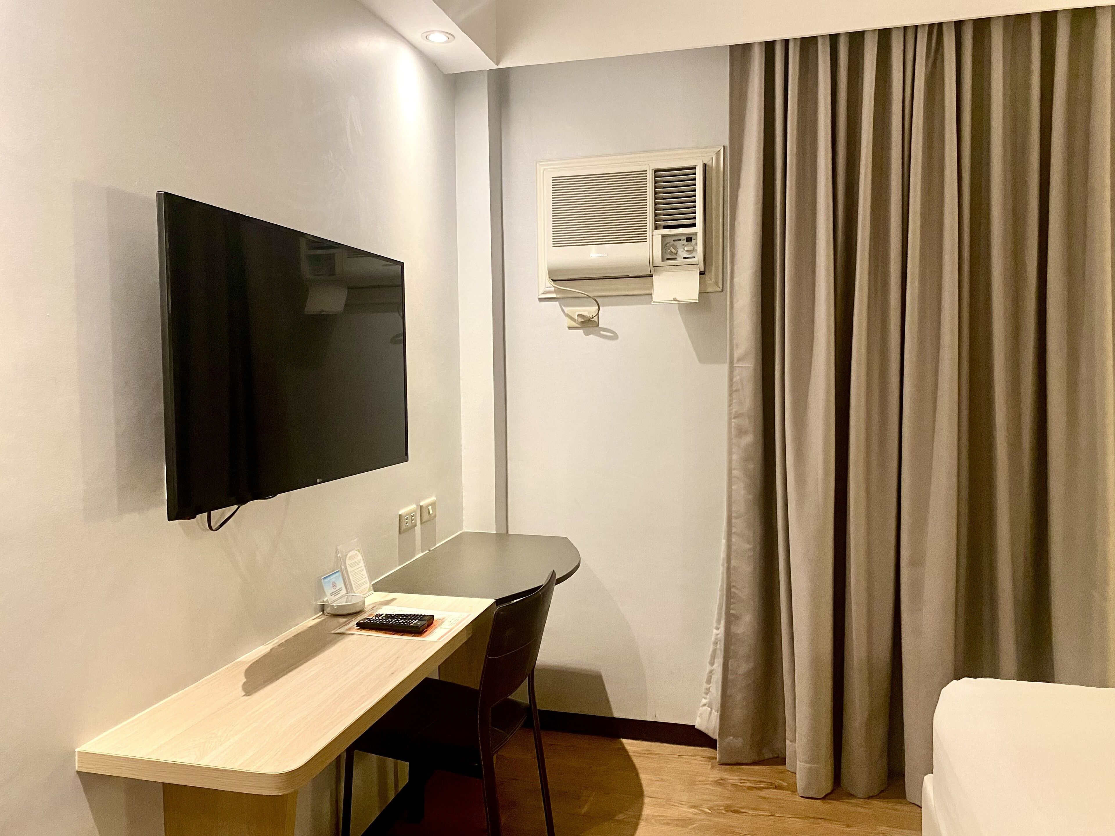 A hotel room with a flat-screen TV mounted on the wall, a desk with a chair, a small table, and a window with curtains.