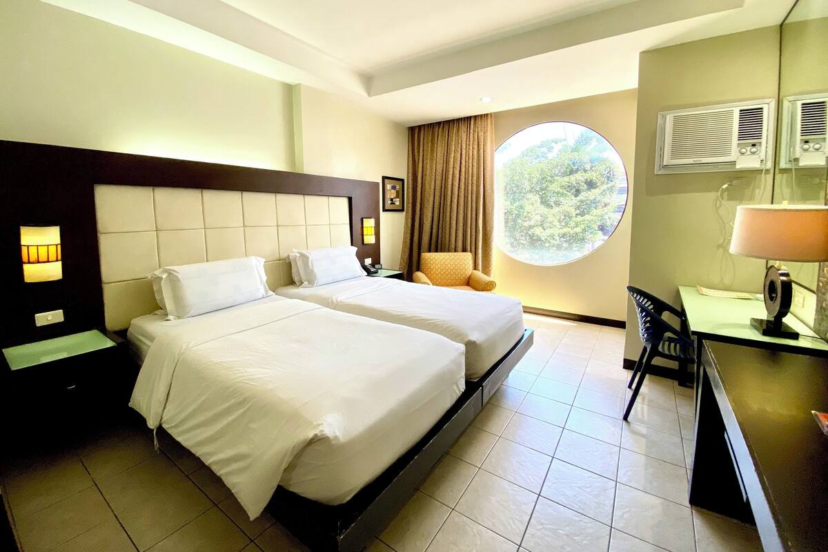 Quarto de hotel com duas camas, uma escrivaninha, uma cadeira, uma janela redonda com vista e uma luminária no teto.