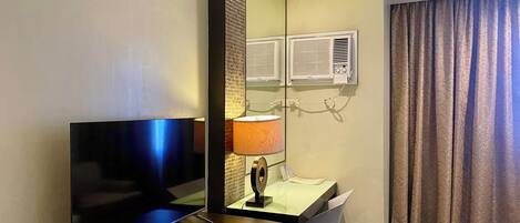 Habitación de hotel con televisión de pantalla plana, un escritorio con lámpara, una silla y un ventanal con cortinas.