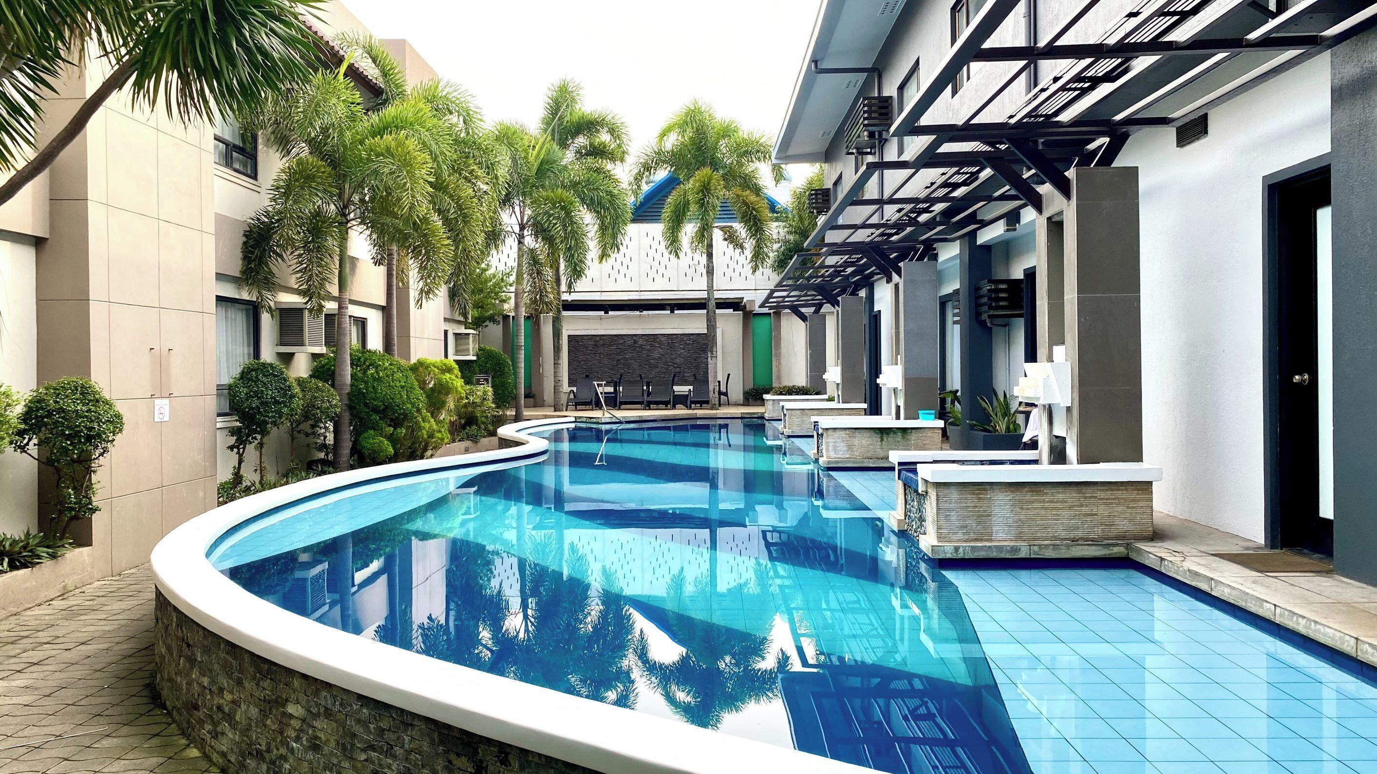 A modern swimming pool with a curved design, surrounded by palm trees and a building with a contemporary architectural style.