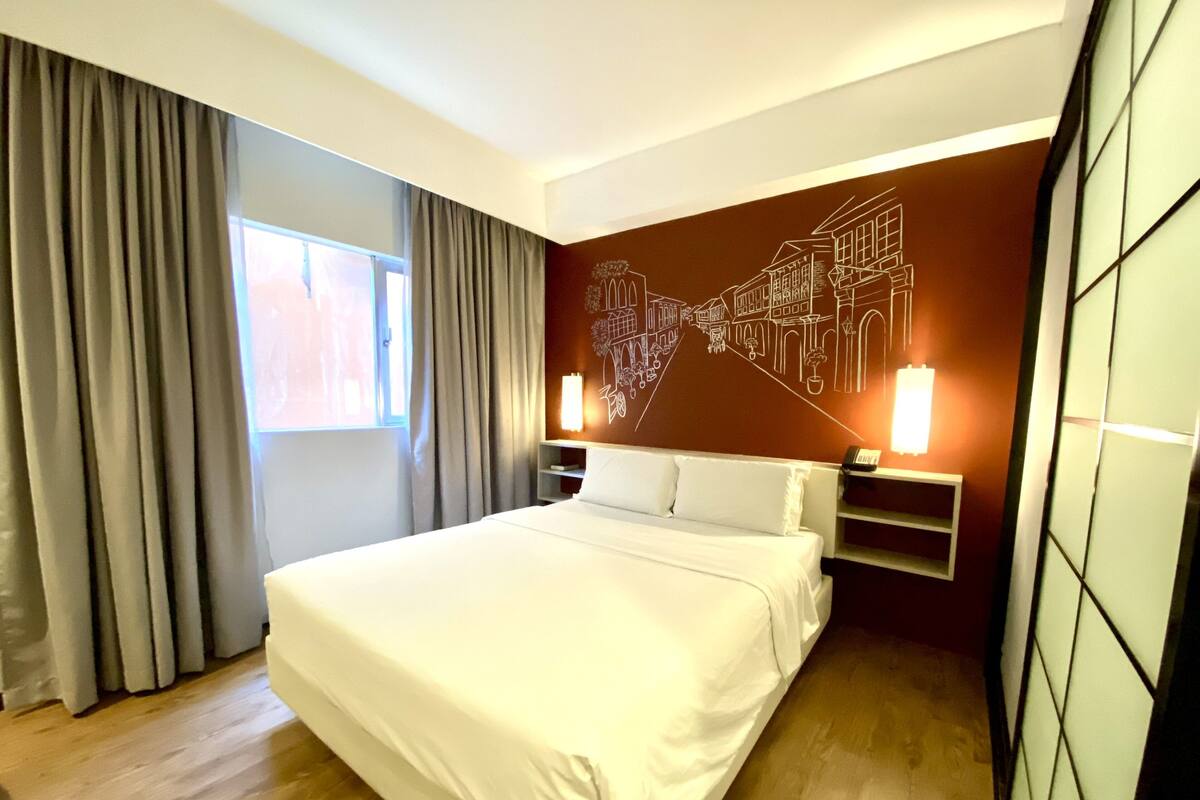 Quarto de hotel moderno com uma cama grande, uma mesa de cabeceira, uma luminária fixada na parede e uma janela com cortinas.