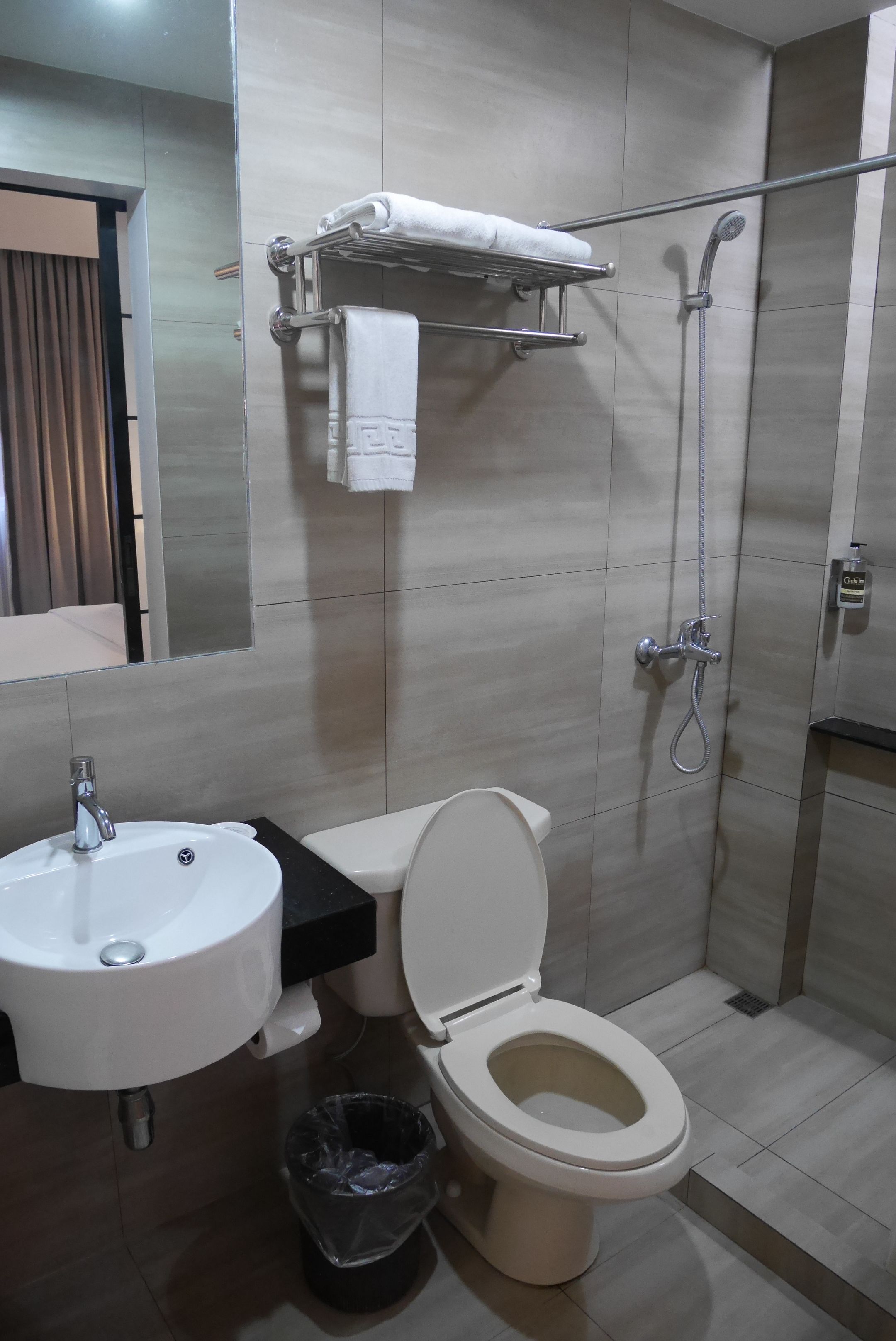 A modern bathroom with a white sink, toilet, and shower. There is a towel rack above the toilet and a trash bin next to it.