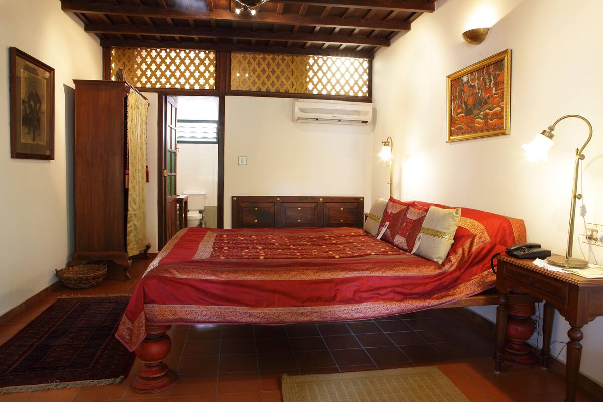 A traditional room with a large bed, a wooden headboard, a desk with a lamp, and a wall-mounted air conditioner.
