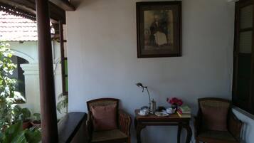A room with two wooden chairs, a small table with a lamp, a framed picture on the wall, and a window with a view of greenery.