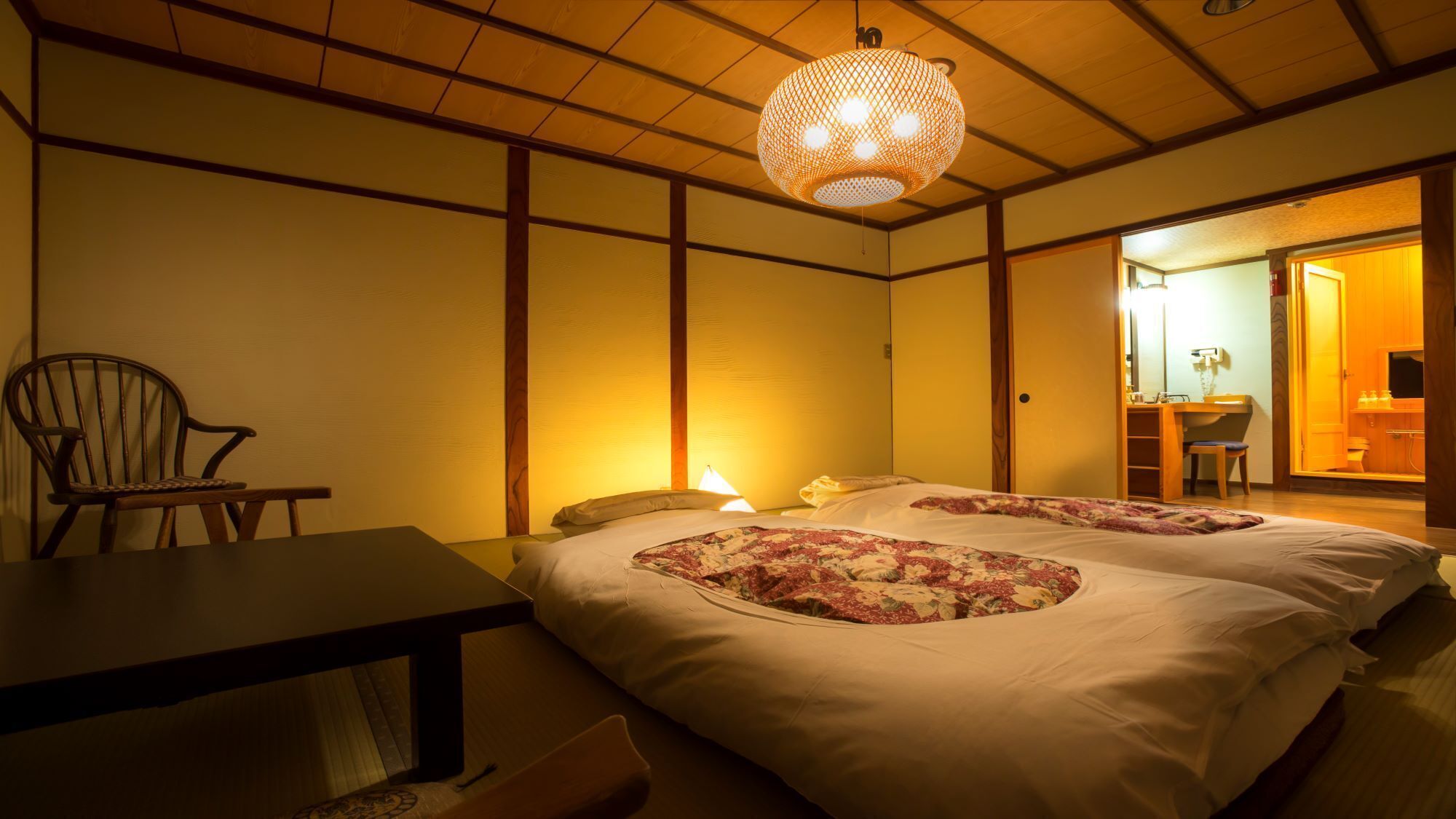 Une pièce de style japonais traditionnel, avec un sol recouvert de tatamis, une basse table en bois, un lit avec une literie à motifs floraux et une lanterne en papier suspendue.