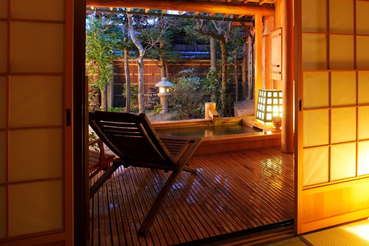 A traditional Japanese room with tatami flooring, a wooden chair, and a view of a garden with a water feature.