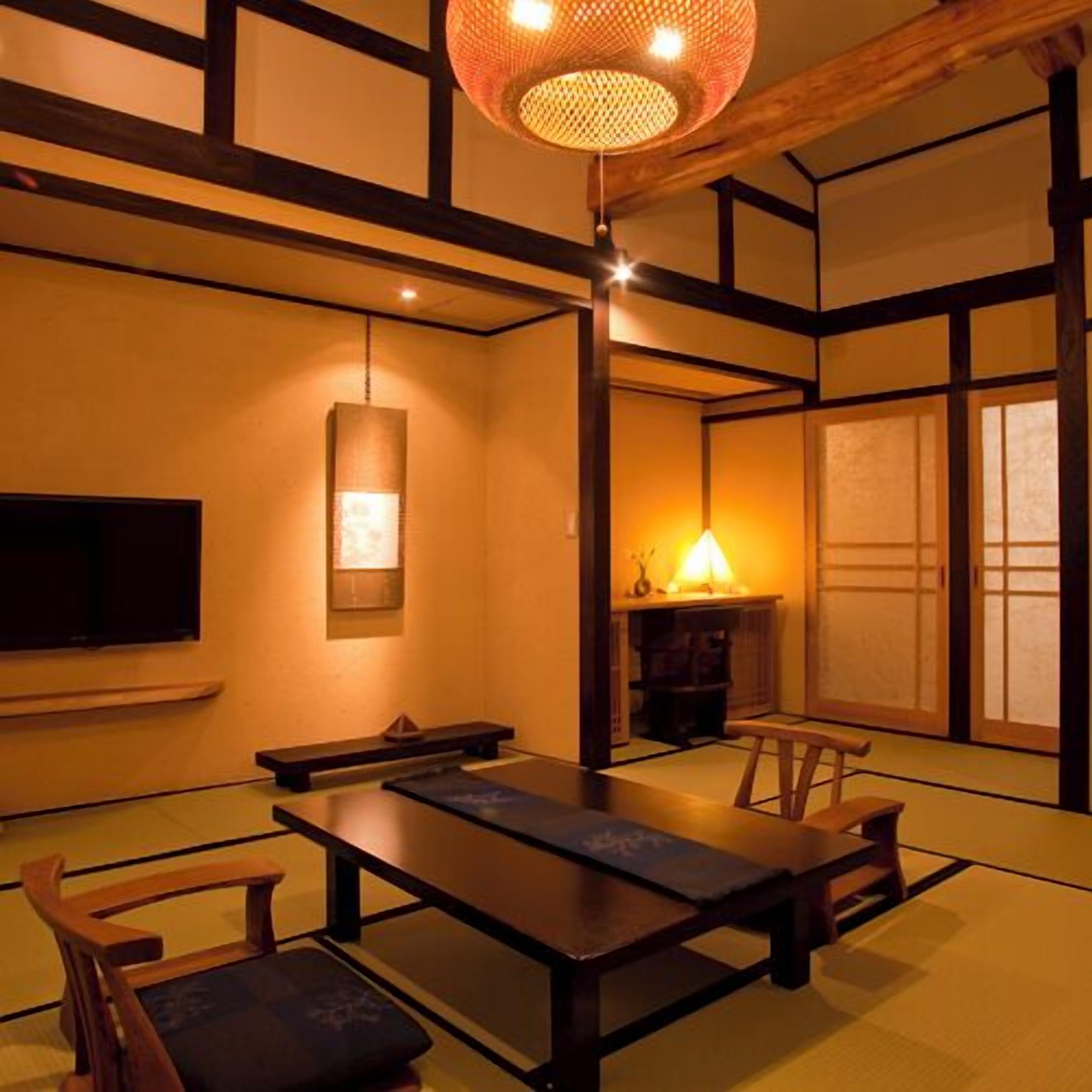 Ein traditioneller japanischer Raum mit Tatami-Boden, einem niedrigen Tisch, Holzstühlen, einem Fernseher und einer Hängelampe.