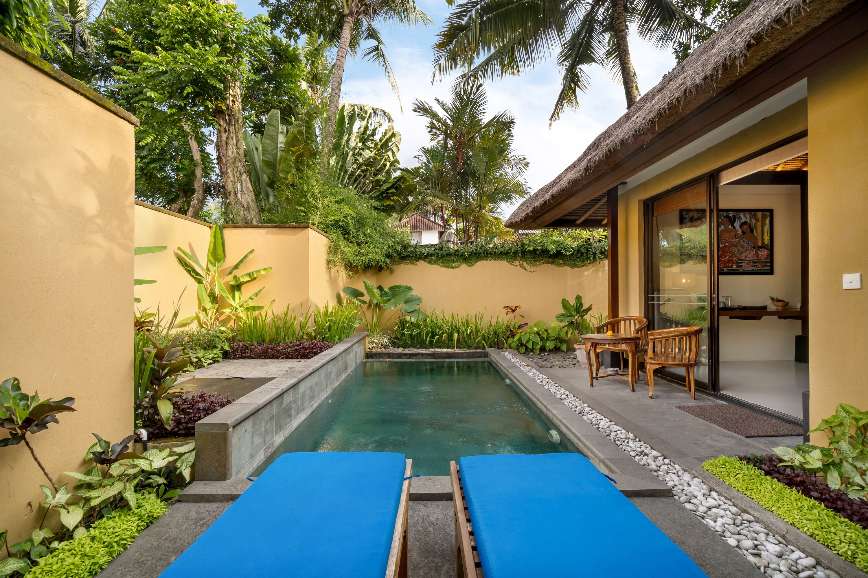 Zona de piscina con sillas de descanso, rodeada de plantas tropicales y una estructura con techo de paja.