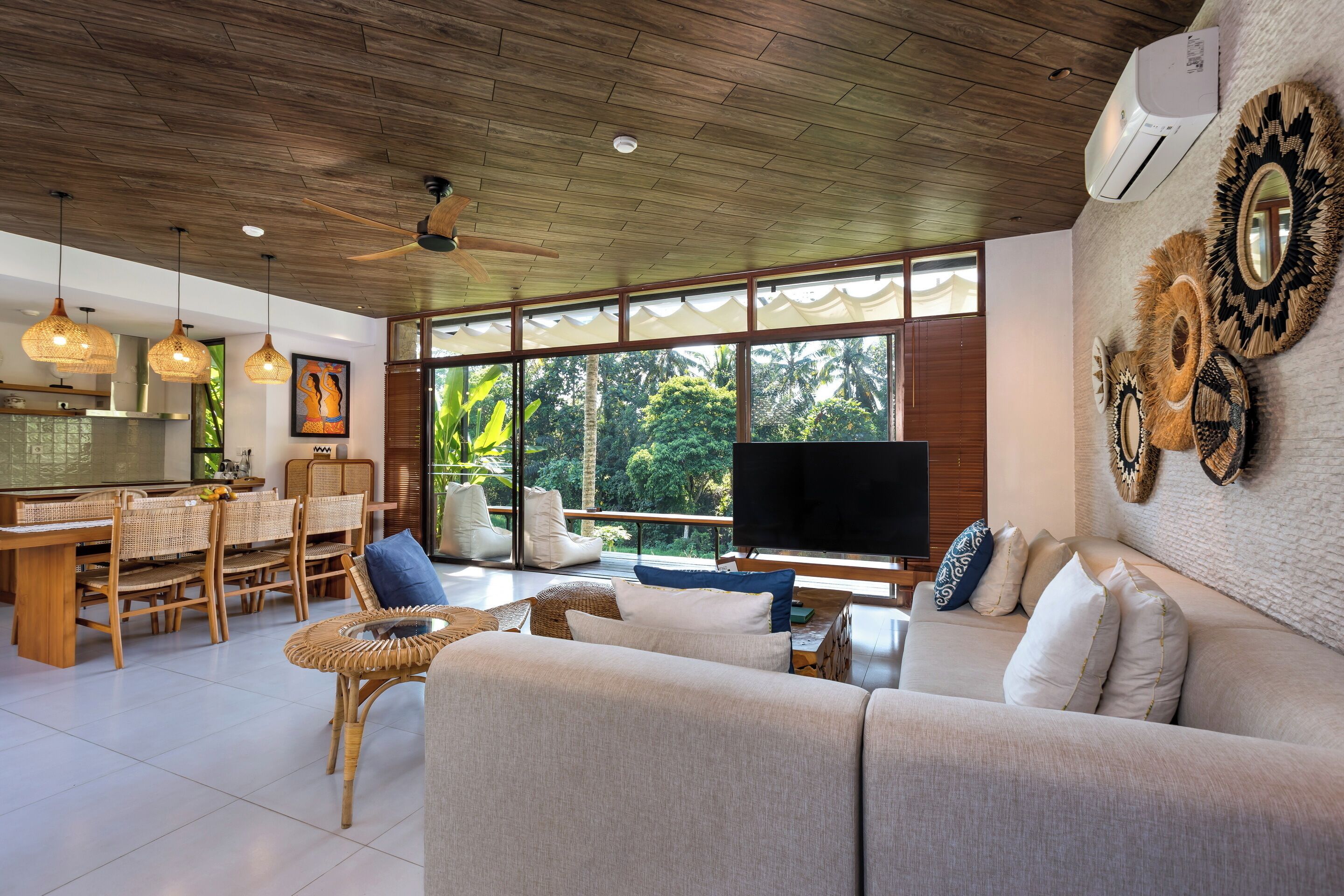 A spacious living area with a sofa, dining table, and chairs, large windows with a view of greenery, and a ceiling fan.
