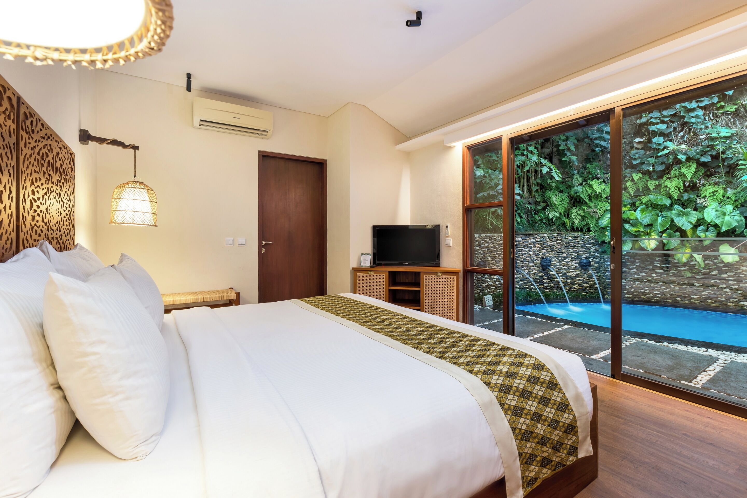A hotel room with a large bed, a television on a wooden stand, and a view of a swimming pool and greenery outside.