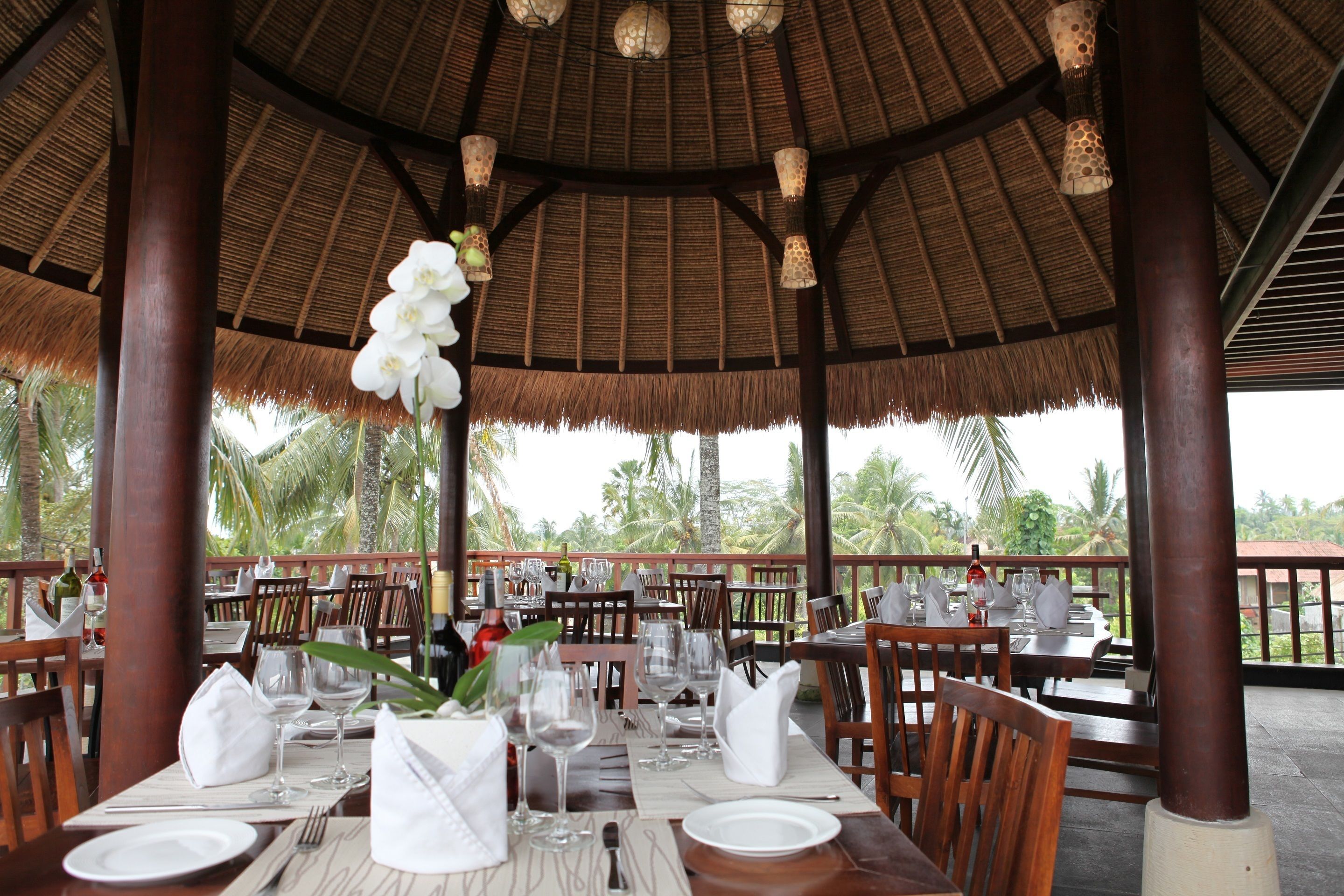 Un restaurant doté d’un toit de chaume, de tables en bois dressées pour le repas et de fleurs blanches suspendues.