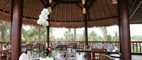 A restaurant with thatched roof, wooden tables set for dining, and hanging white flowers.