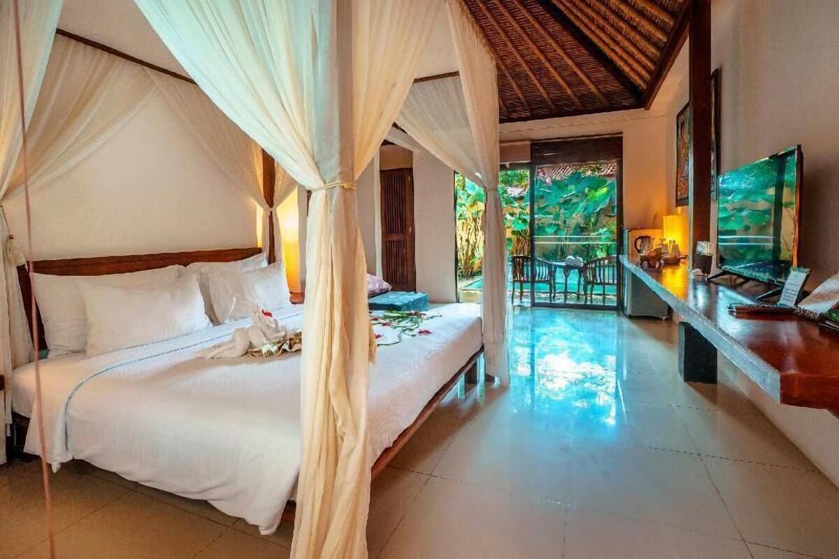 A four-poster bed with a canopy, a television, and a desk with a chair in a room with a tiled floor and a view of greenery outside.