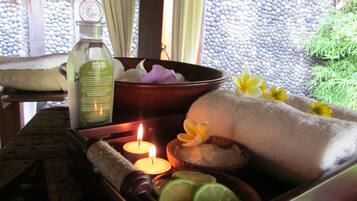 A spa setting with a bottle of Minyak Zaitun oil, a bowl of flowers, a lit candle, and a towel on a table.