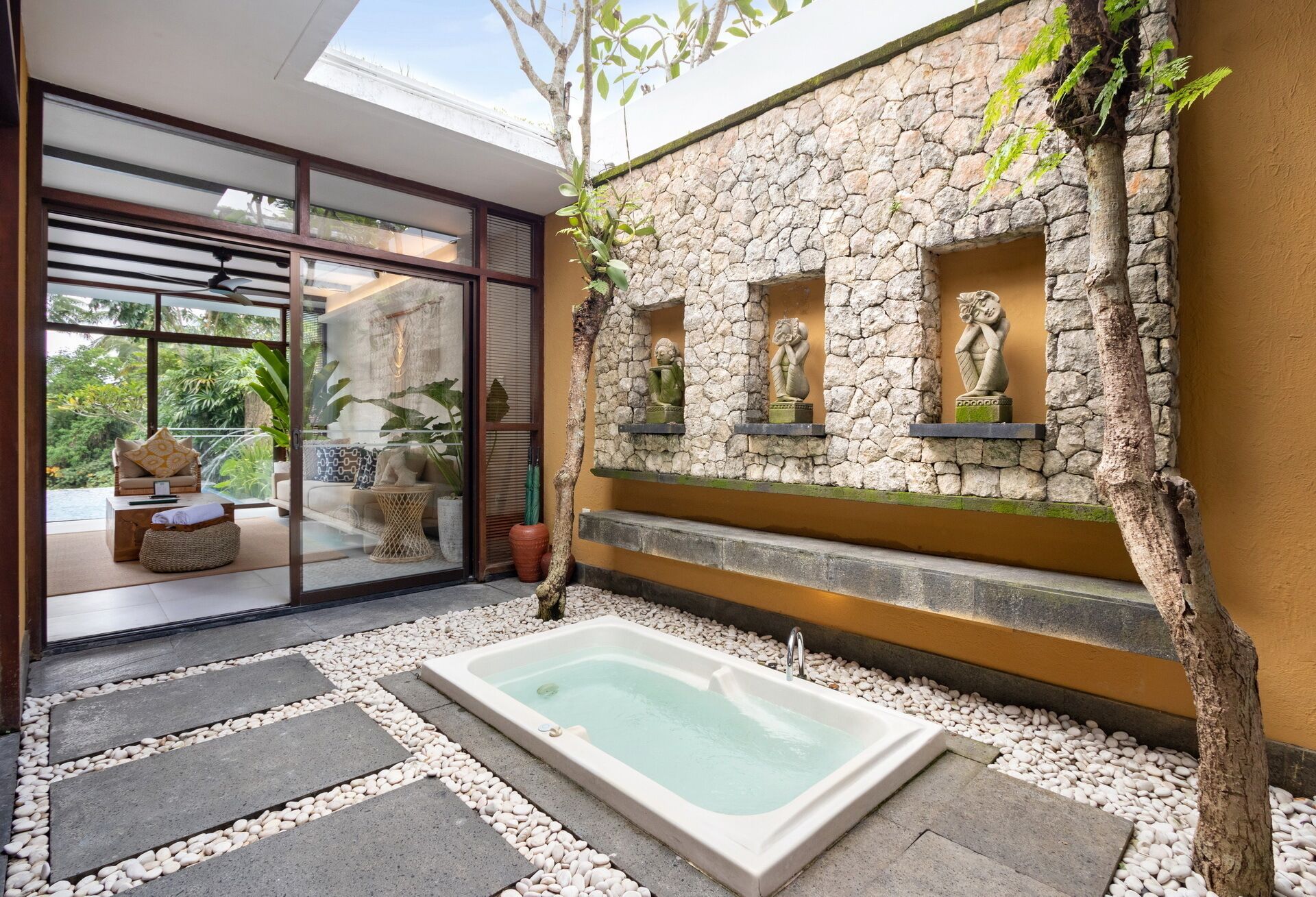Un espace de détente avec un jacuzzi, un mur de pierre orné de statues et une porte vitrée donnant sur un coin salon avec vue sur la verdure.