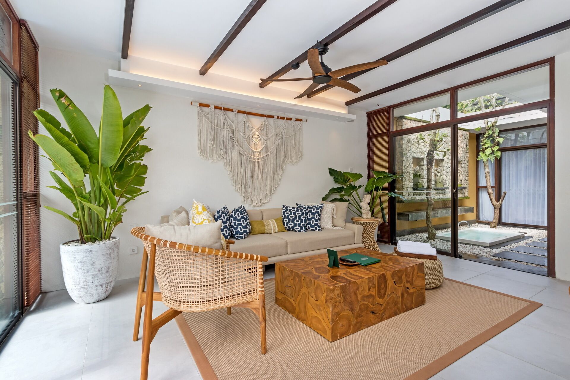 Una sala de estar moderna con sofá, mesa de centro y silla de mimbre. Hay una planta grande, un tapiz de macramé y vistas a un jardín a través de una puerta de cristal.