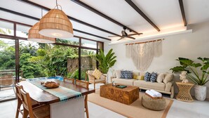 Ampla sala de estar com mesa de jantar, sofá e grandes janelas com vista para um jardim exuberante.