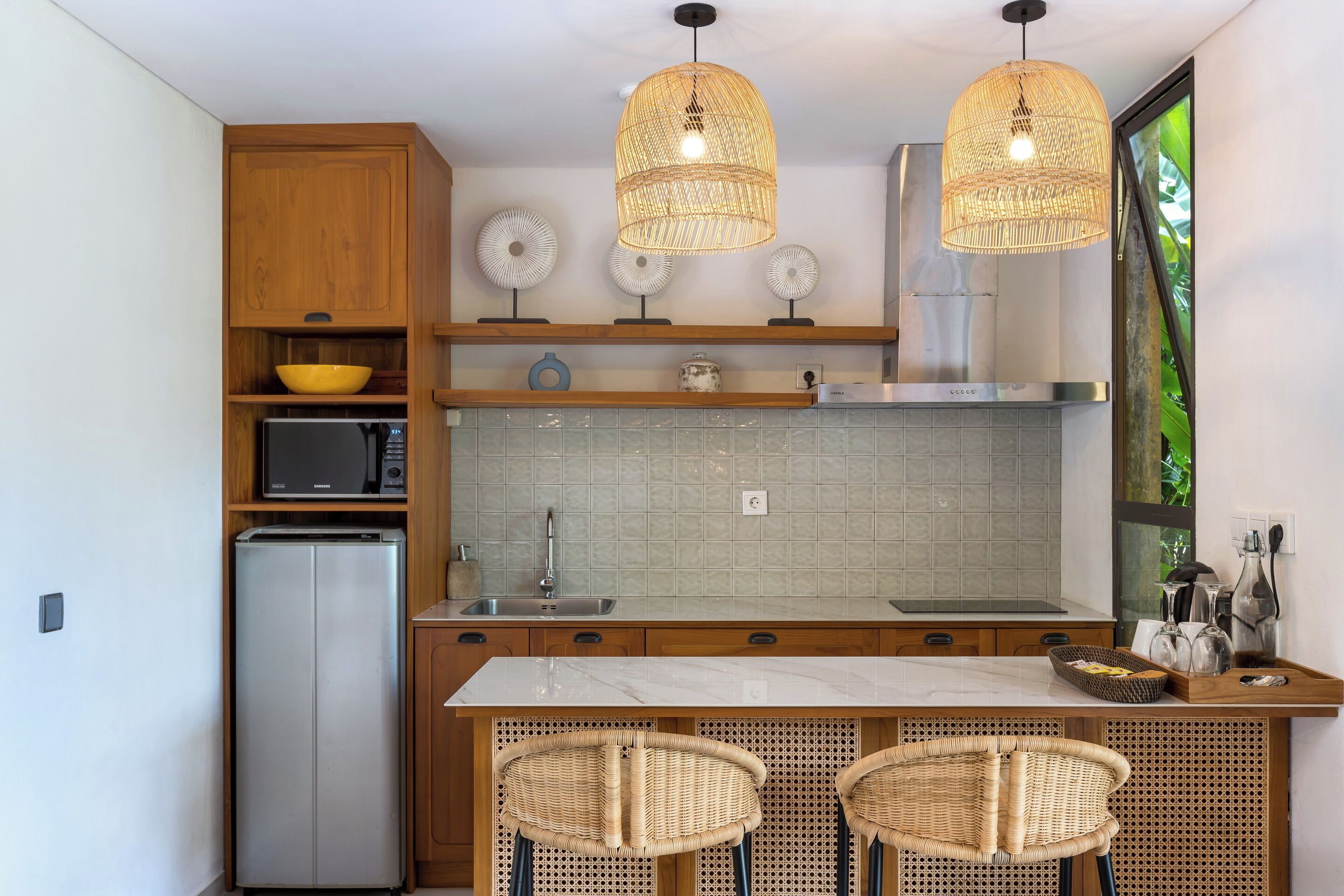 A compact kitchen with wooden cabinets, a built-in microwave, a stainless steel refrigerator, a marble countertop, two wicker chairs, and a hanging light fixture.