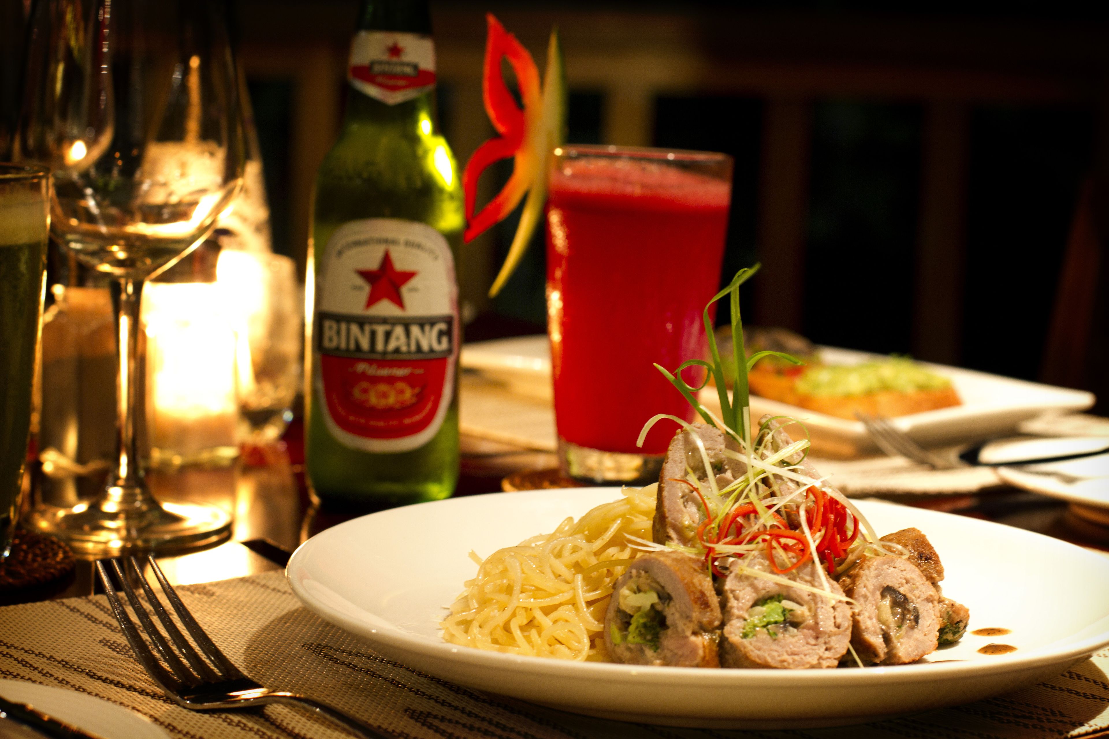 A dining table with plates of food, a bottle of Bintang beer, a glass of red drink, and a bottle of wine.