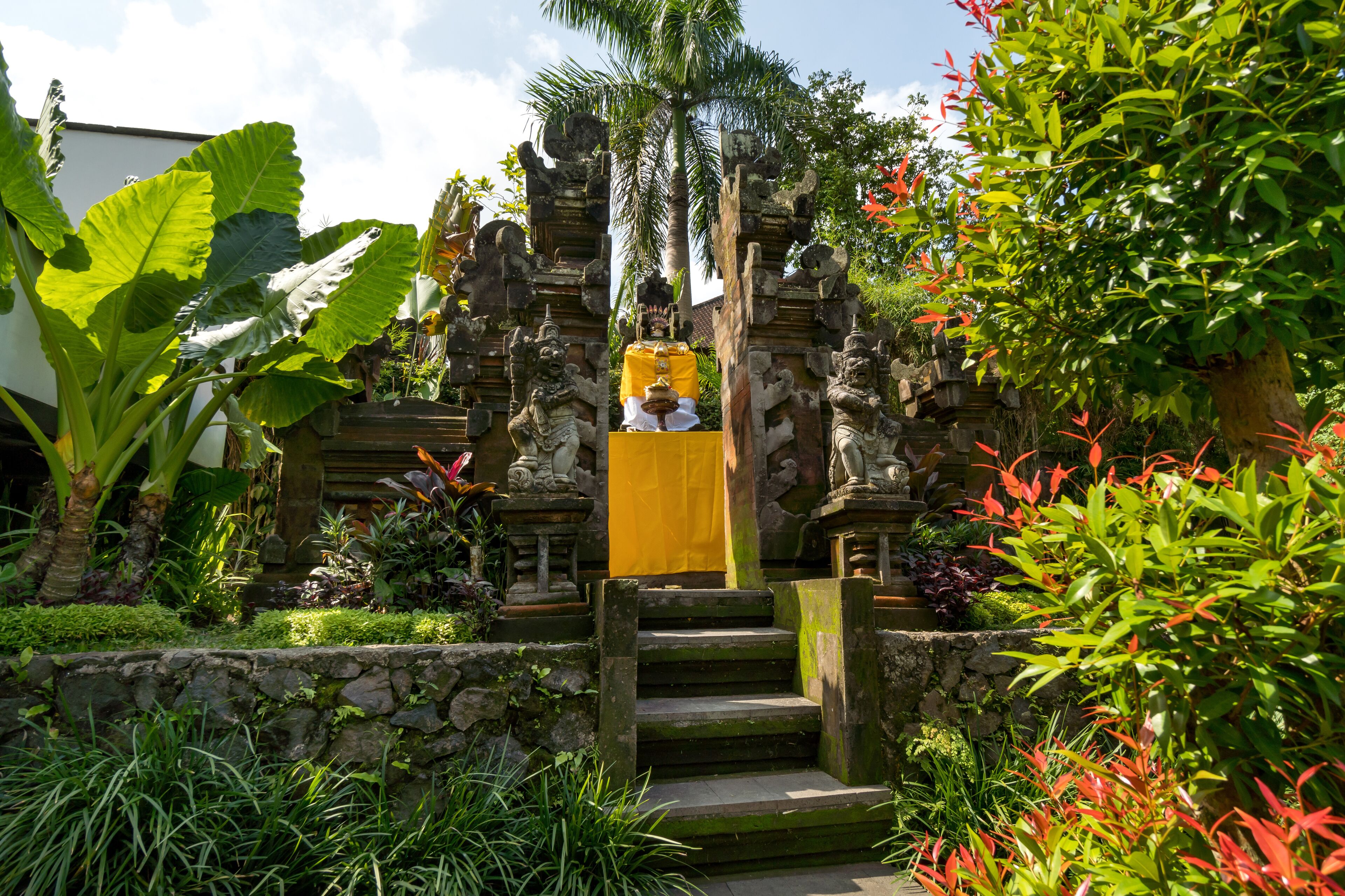 A traditional Balinese temple with stone carvings, a yellow cloth, and lush greenery.