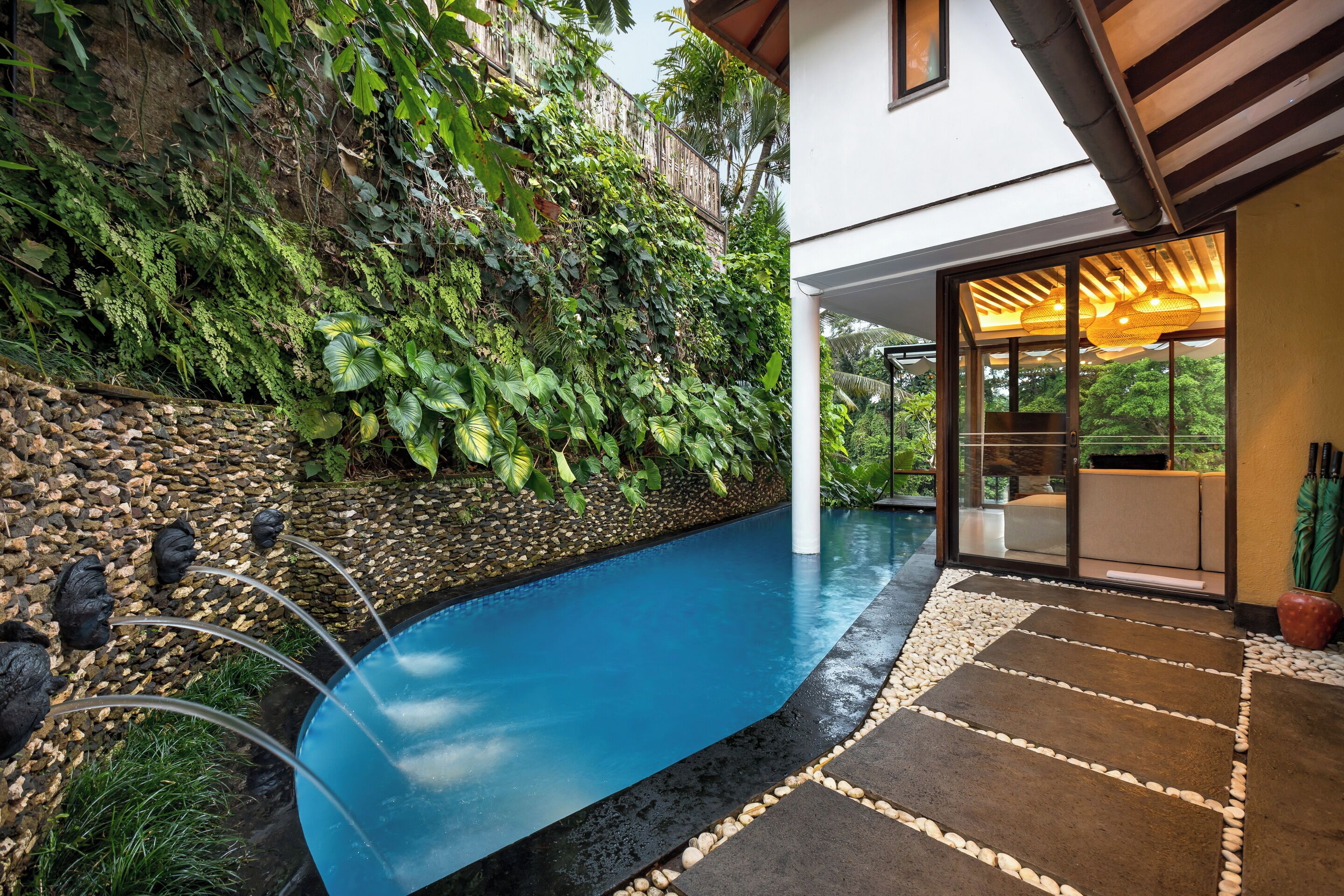 A modern outdoor pool area with a stone-lined edge, a waterfall feature, and lush greenery surrounding the space.