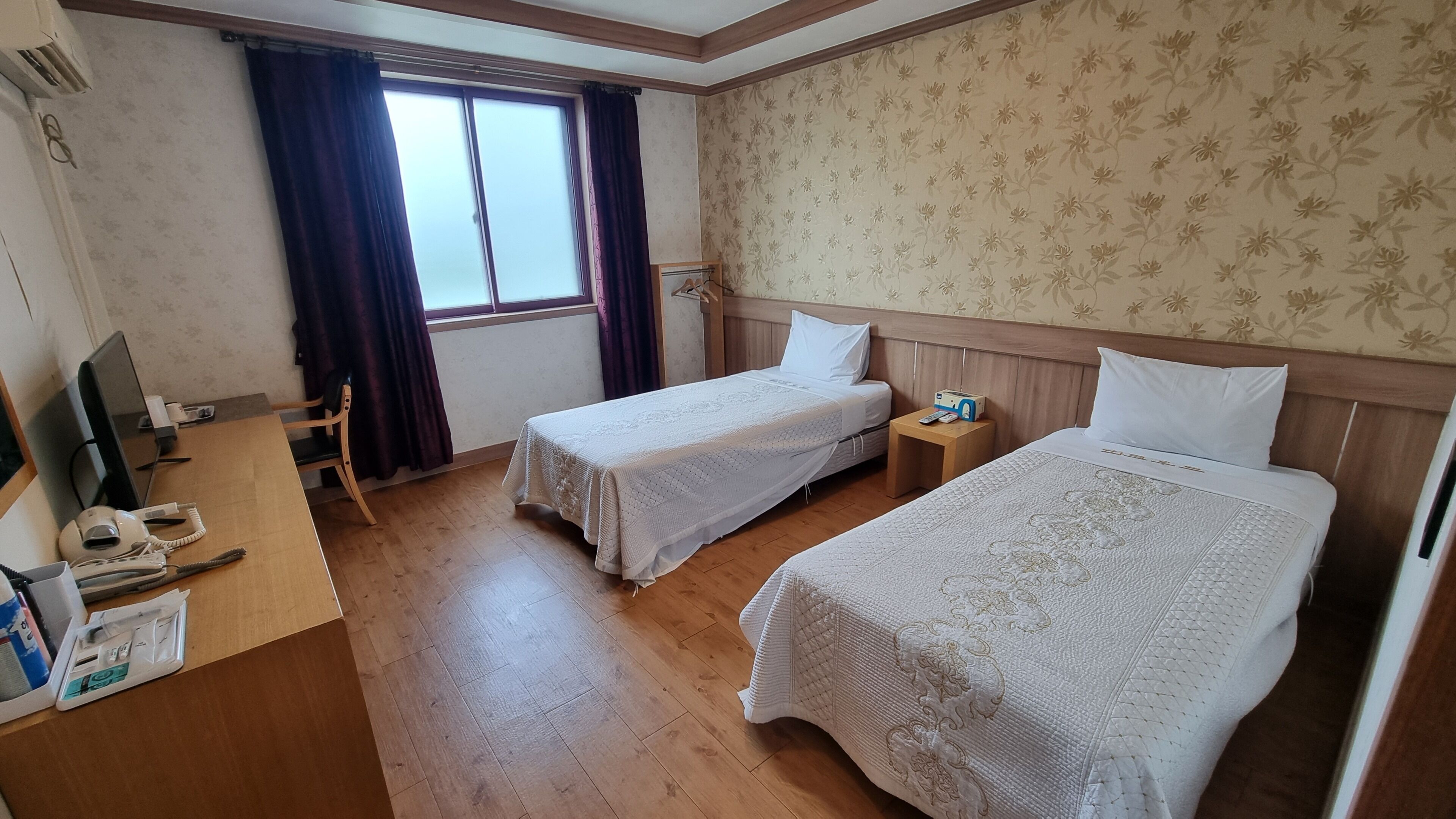 Een hotelkamer met twee bedden, een bureau met computer en telefoon, een raam met gordijnen en een houten vloer.