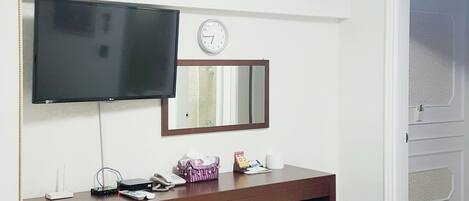 A hotel room with a wooden desk, a flat-screen TV, a clock, a mirror, a small refrigerator, a trash bin, and a door.