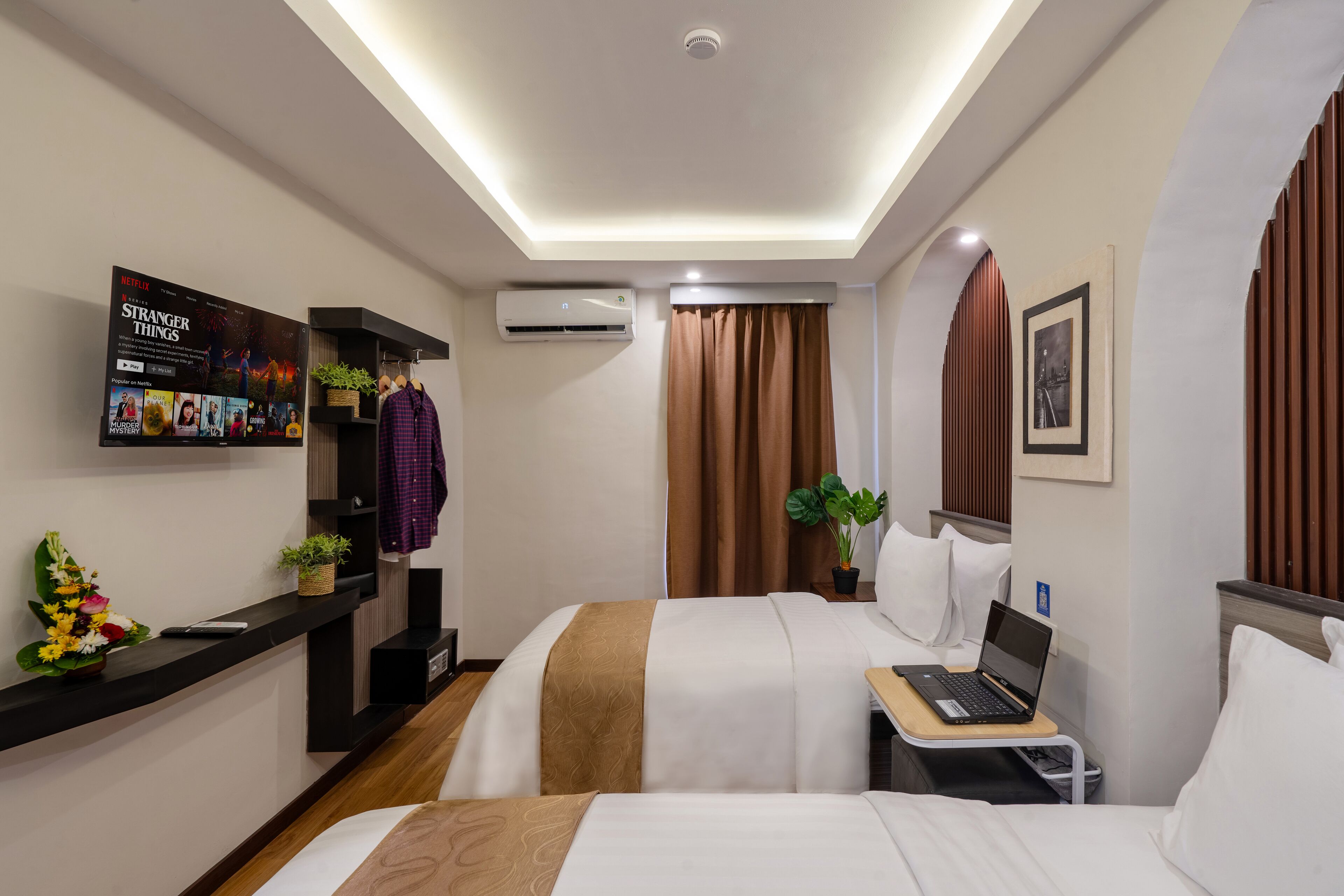 Habitación de hotel con dos camas, un escritorio con una computadora portátil, un equipo de aire acondicionado, un televisor mostrando 'Stranger Things' y una repisa de pared con una planta y un objeto decorativo.