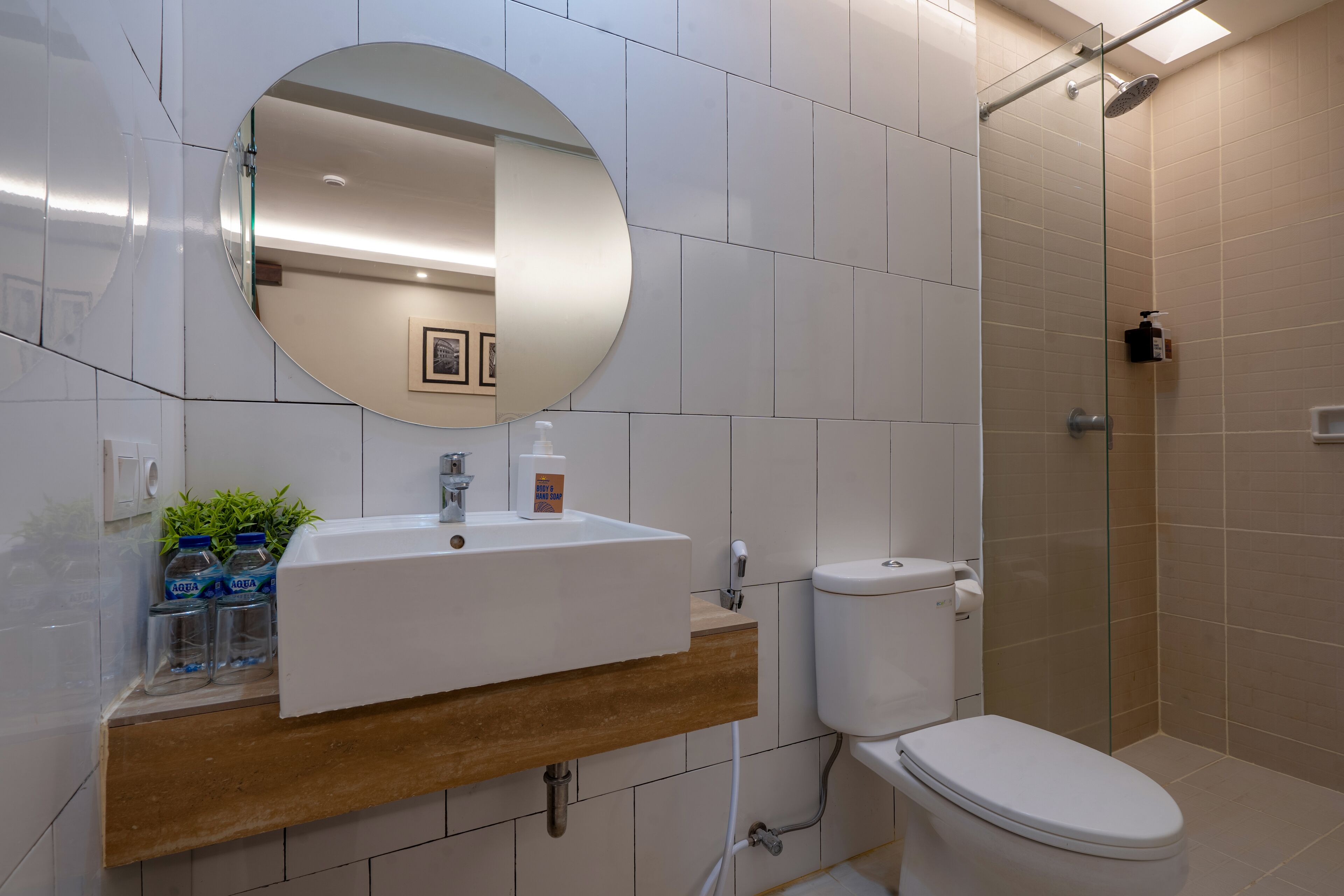 A modern bathroom with a white sink, mirror, and toilet. There is a shower area with a glass door and a small shelf with toiletries.