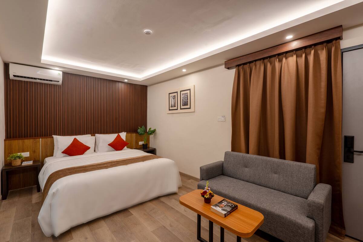 A hotel room with a bed, a grey sofa, a wooden coffee table, and a wall-mounted air conditioner.