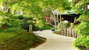Un sendero de madera serpentea por un exuberante jardín con un edificio tradicional japonés al fondo.