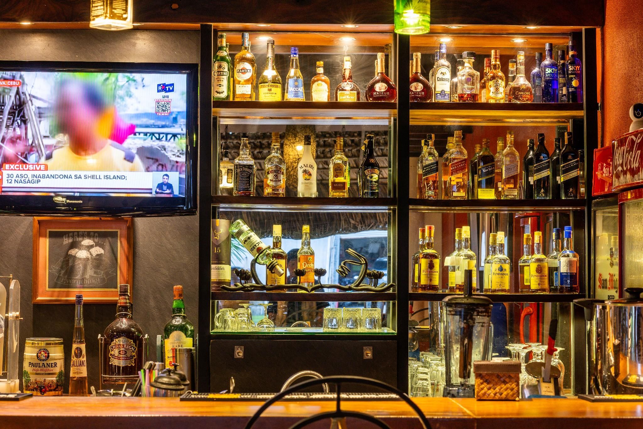 Zona de bar con varios estantes llenos de botellas de licores, una pantalla de televisión y un cuadro en la pared.