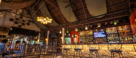 A bar area with multiple seating arrangements, a television screen, and a variety of bottles on shelves.
